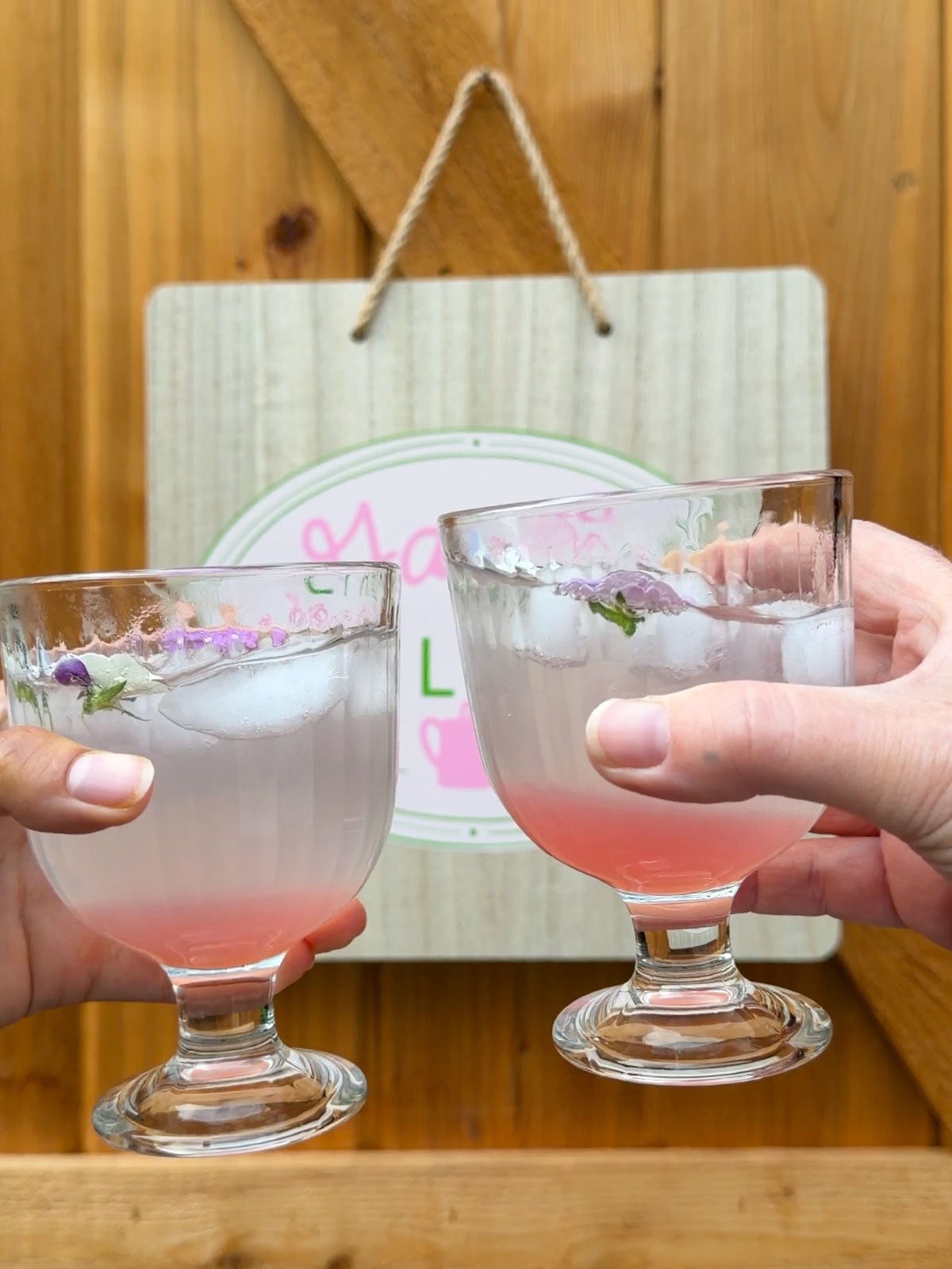 Two hands raising glasses of rhubarb soda in a toast in front of the Garden Club sign.