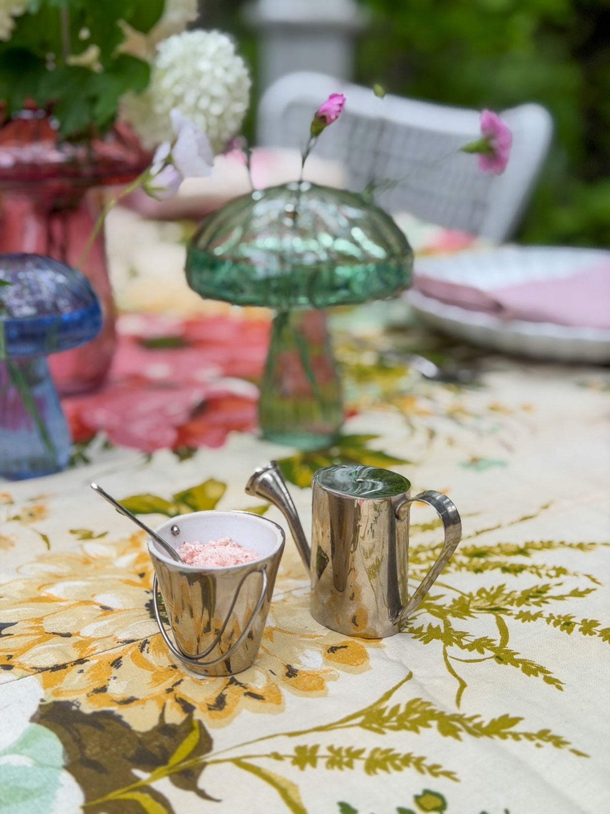 Miniature watering can and bucket salt and pepper shakers on floral tablecloth with glass mushroom vase in background.