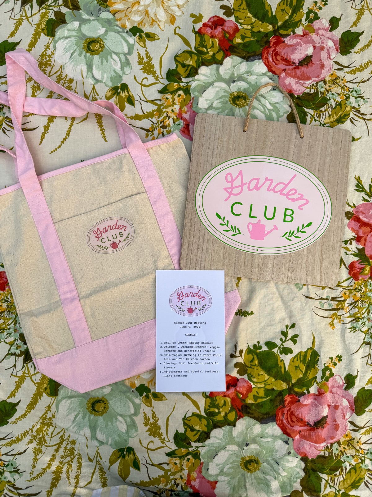 Garden Club tote bag with logo, hanging sign, and printed agenda card arranged on vintage floral tablecloth.