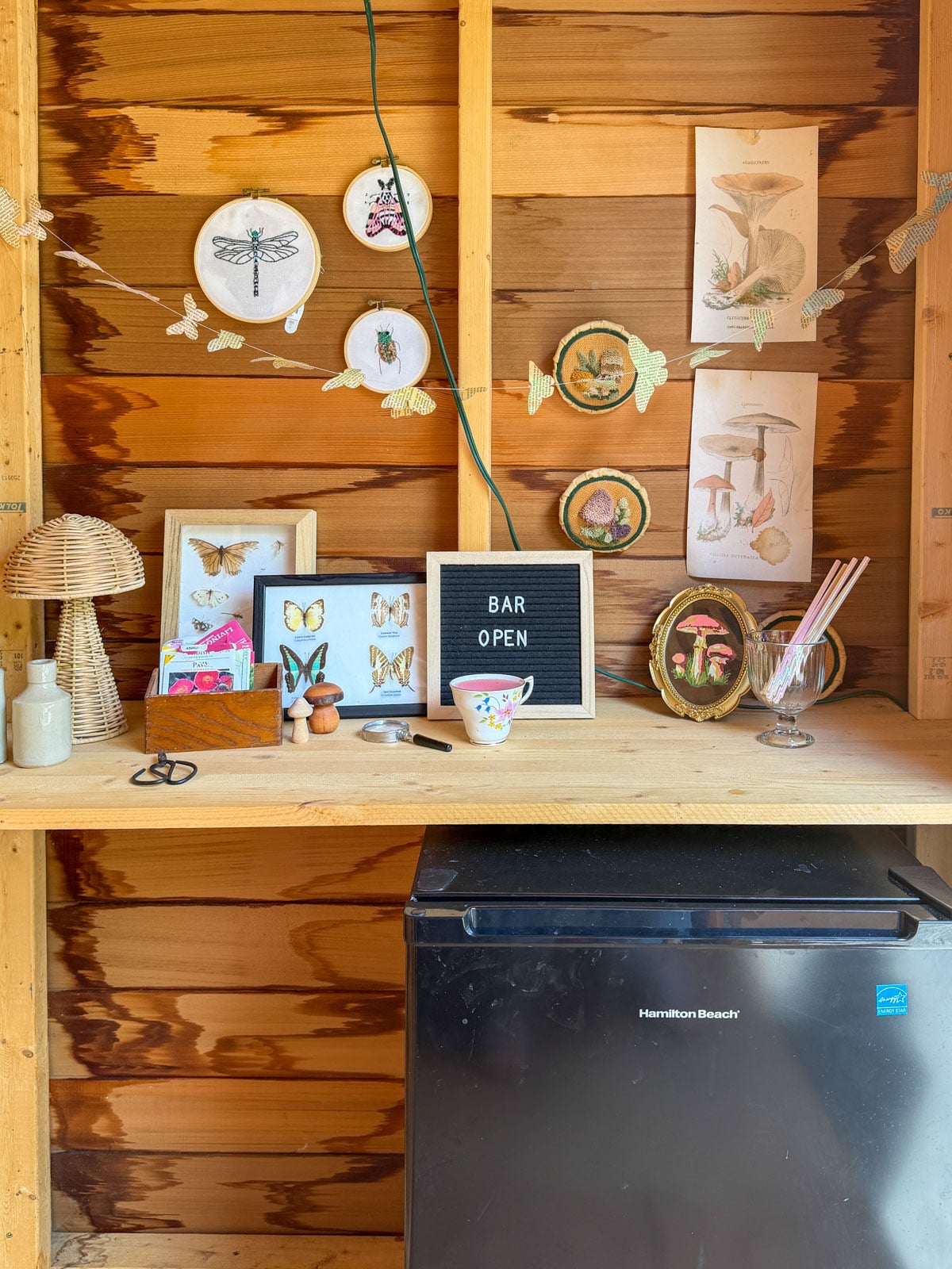 Garden shed bar setup with Bar Open letter board, butterfly embroidery hoops, mushroom botanical prints, seed packet garland, and mini bar fridge.