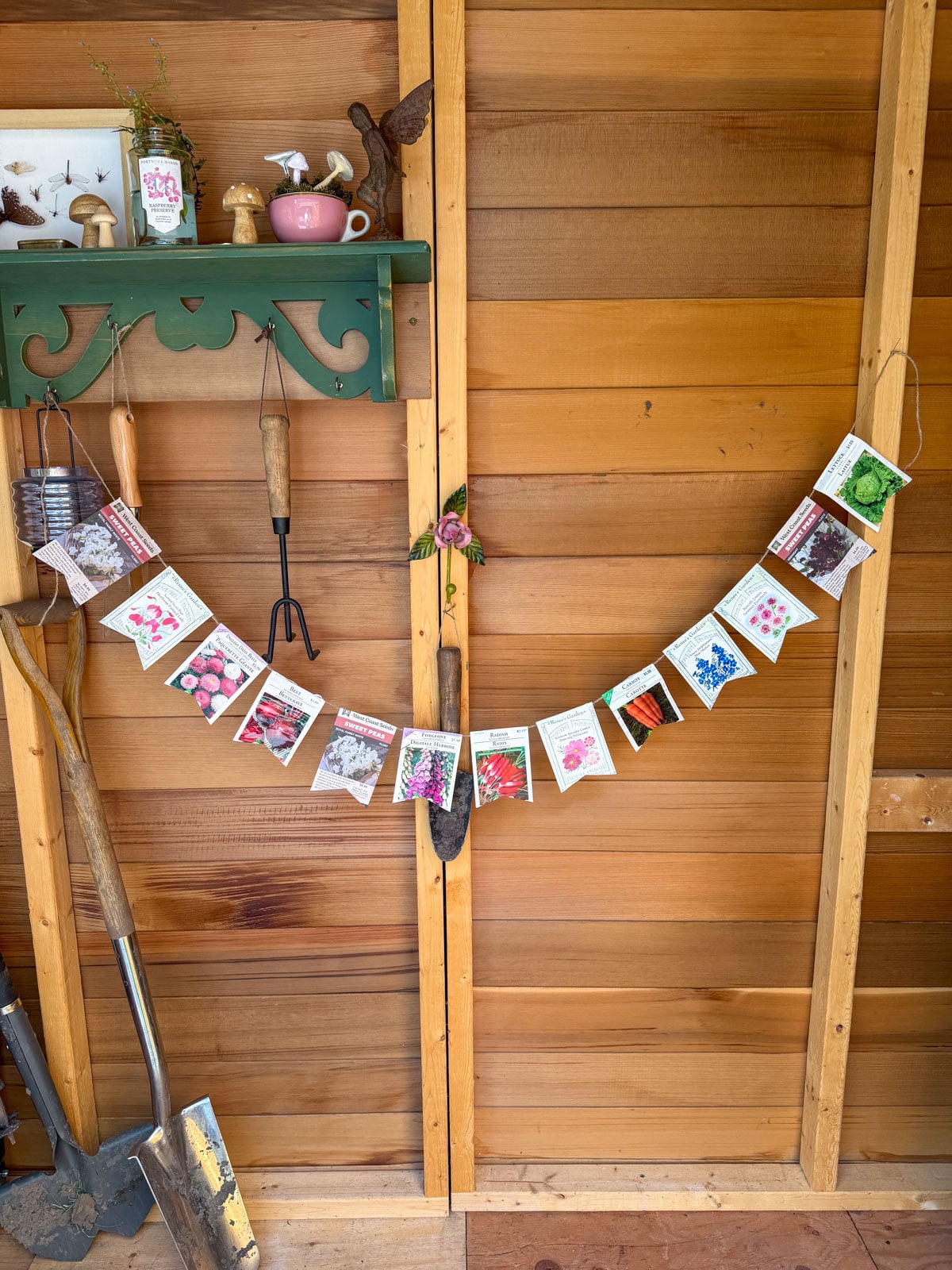 Seed packet bunting garland draped across garden shed door with garden tools and decorative shelf.