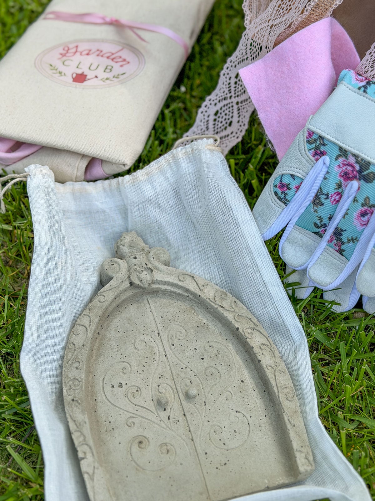 Handmade concrete fairy door garden ornament with Garden Club tote bag and floral garden gloves on grass at plant exchange.