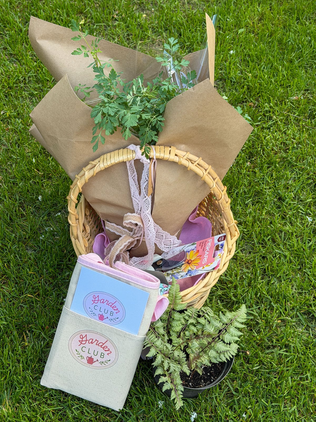 Garden party plant exchange gifts on lawn including thrifted basket with lace bow, Garden Club tote bag, wrapped perennial plant, and floral garden gloves.
