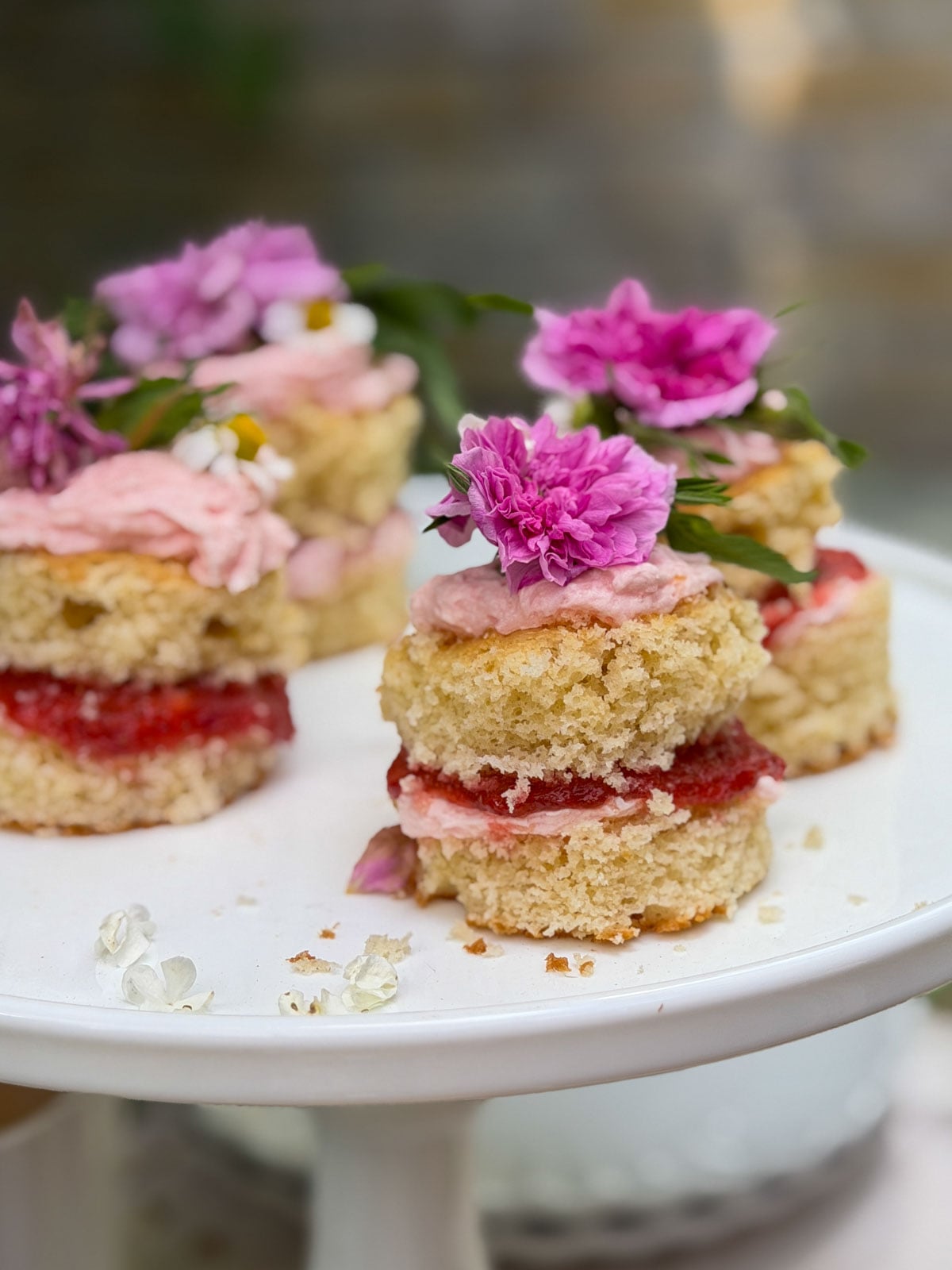 Mini individual vanilla cakes with strawberry rhubarb compote filling, pink frosting, and fresh garden roses and carnations on white cake stand.