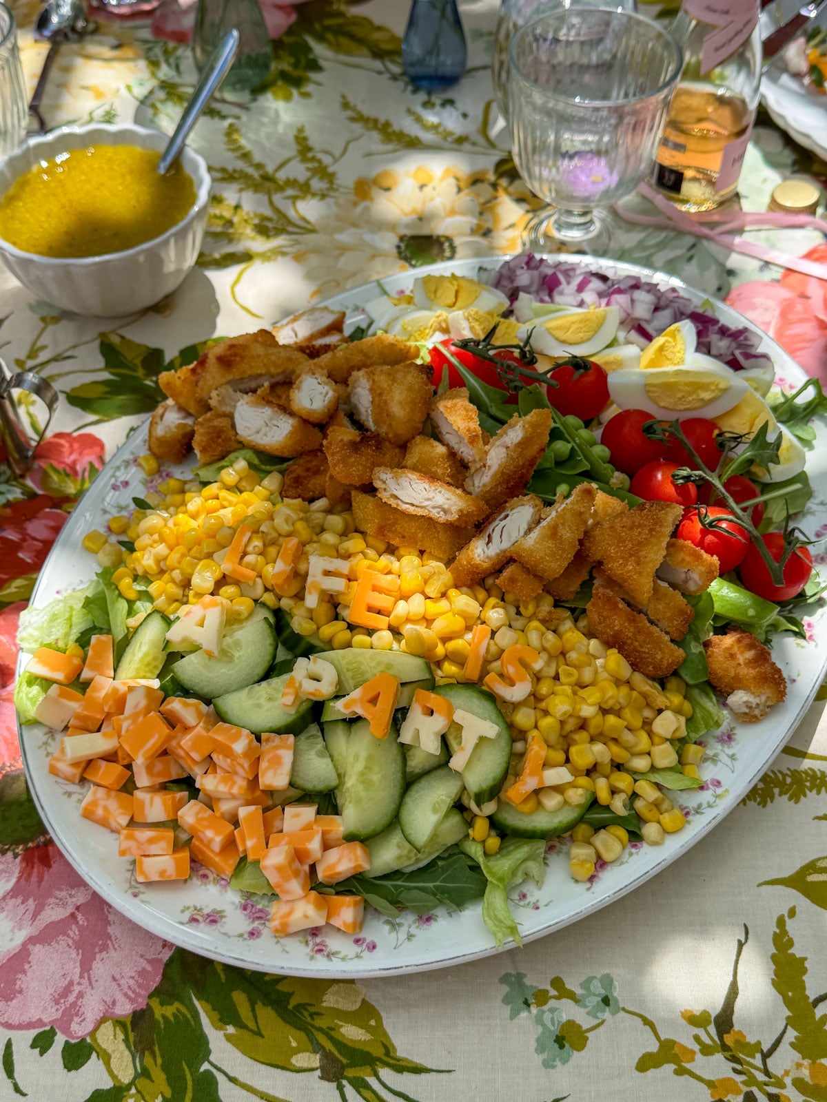 Garden salad with Life is a Party spelled in marble cheese letters, sliced chicken fingers, corn, cucumber, cherry tomatoes, and hard boiled eggs on vintage china platter.