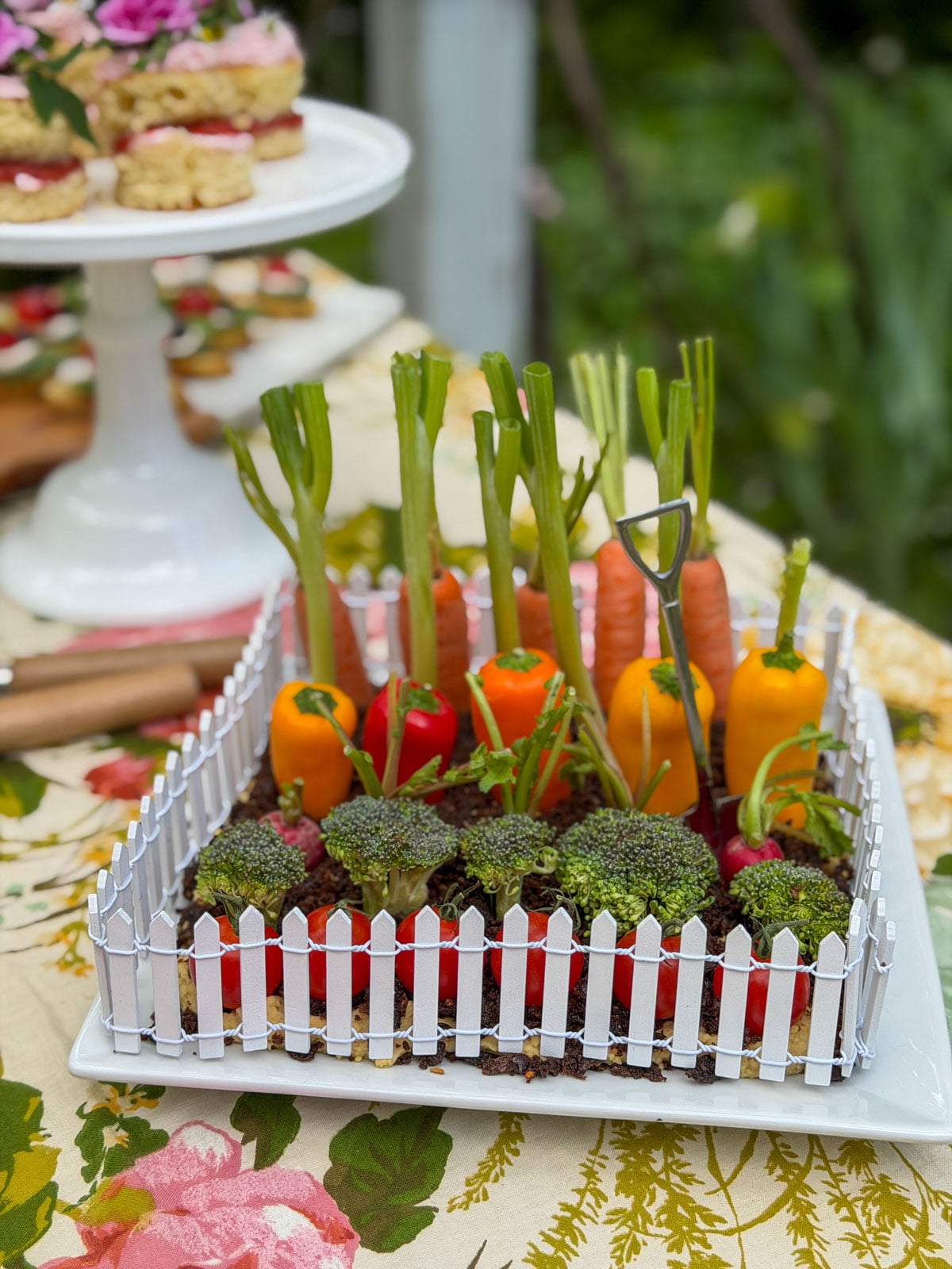 Vegetable garden hummus dip with picket fence on garden party table beside mini vanilla cakes on white cake stand.