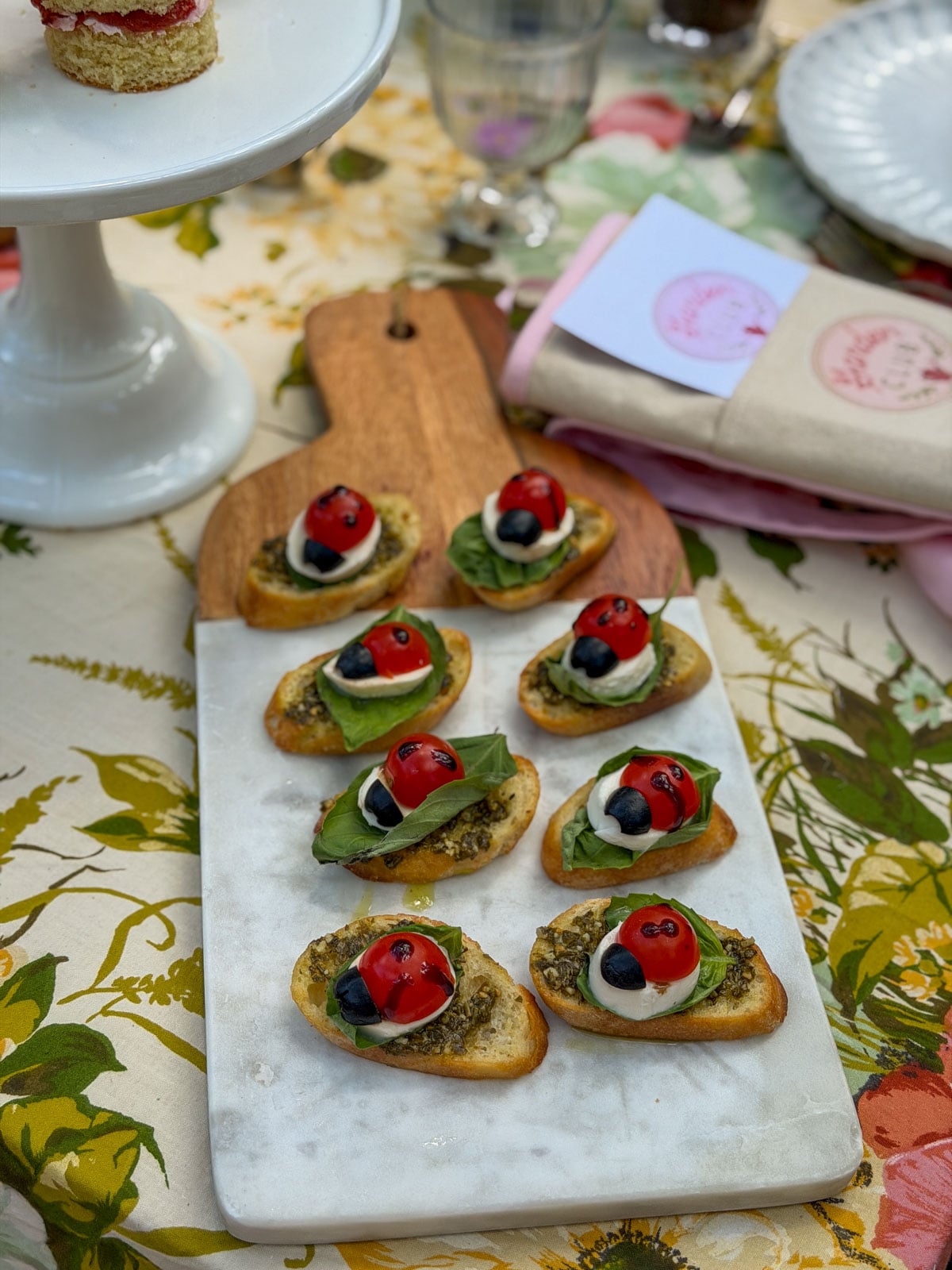Ladybug caprese bruschetta on marble board with cherry tomato ladybugs, fresh mozzarella, basil leaves, pesto, and balsamic glaze on garlic crostini.