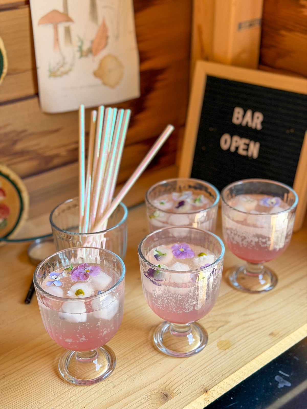 Four glasses of rhubarb soda with floating edible pansies on garden shed bar shelf with Bar Open sign.