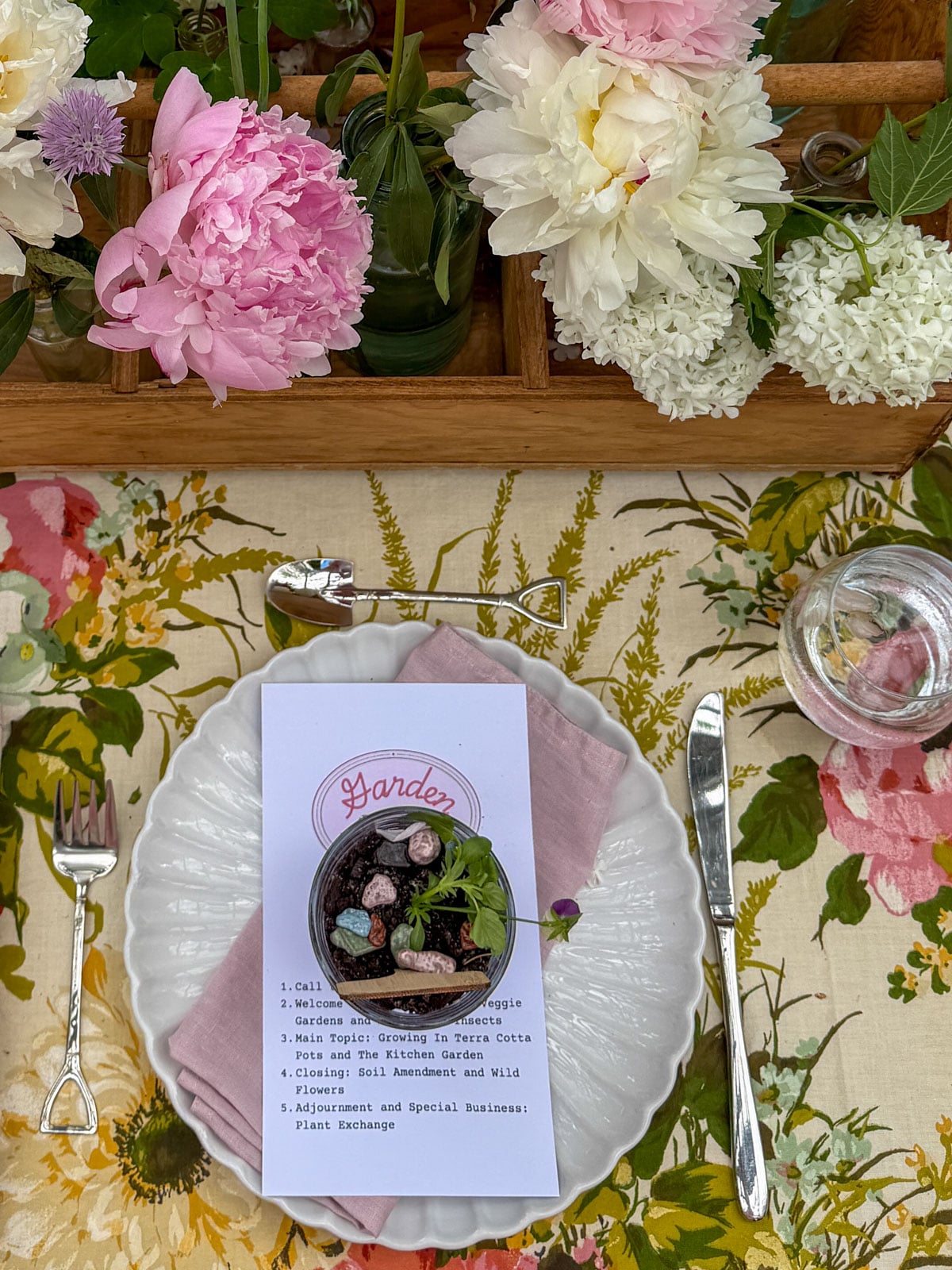 Overhead garden party place setting with white scalloped plate, pink linen napkin, shovel fork, pitchfork knife, Garden Club menu card, and dirt pudding placecard beside a wooden caddy filled with peonies.