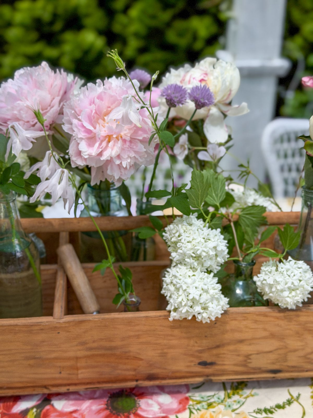 Vintage wooden tool caddy filled with glass bottle vases holding pink peonies, white hydrangeas, and chive blossoms as garden party centerpiece.