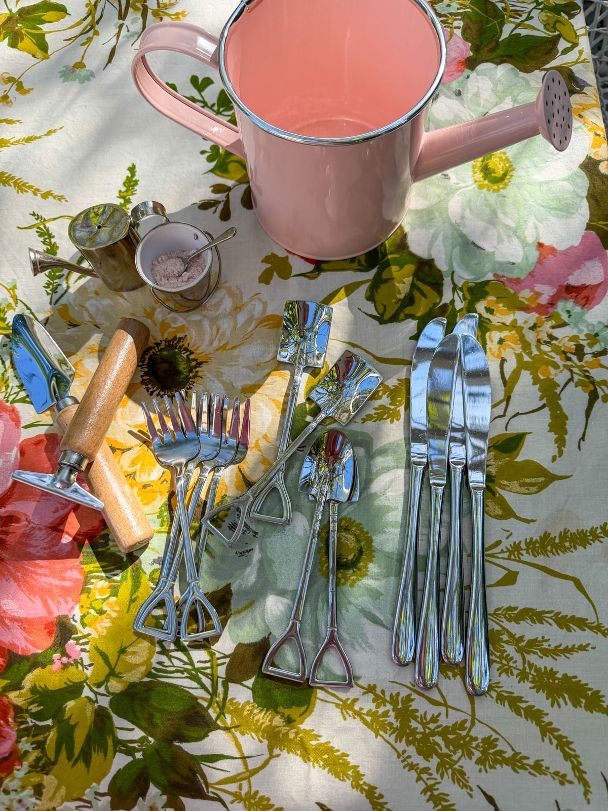Shovel spoons, pitchfork forks, garden trowel and rake salad servers, pink watering can water pitcher, and miniature watering can salt and pepper shakers on vintage floral tablecloth.