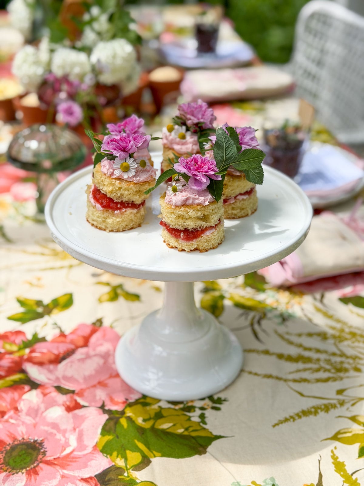 Mini individual vanilla cakes with strawberry rhubarb compote and pink frosting topped with fresh garden roses, daisies, and chive blossoms on white cake stand.