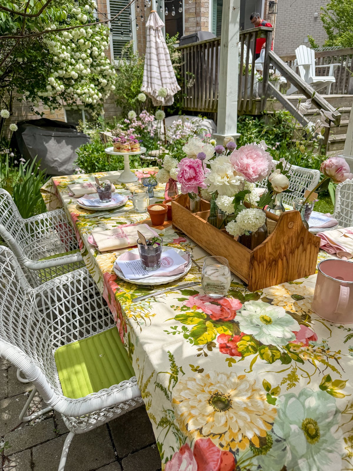 Garden party table set under white pergola with vintage floral tablecloth, pink peony centerpiece in wooden tool caddy, white wicker chairs, and climbing white roses in background.