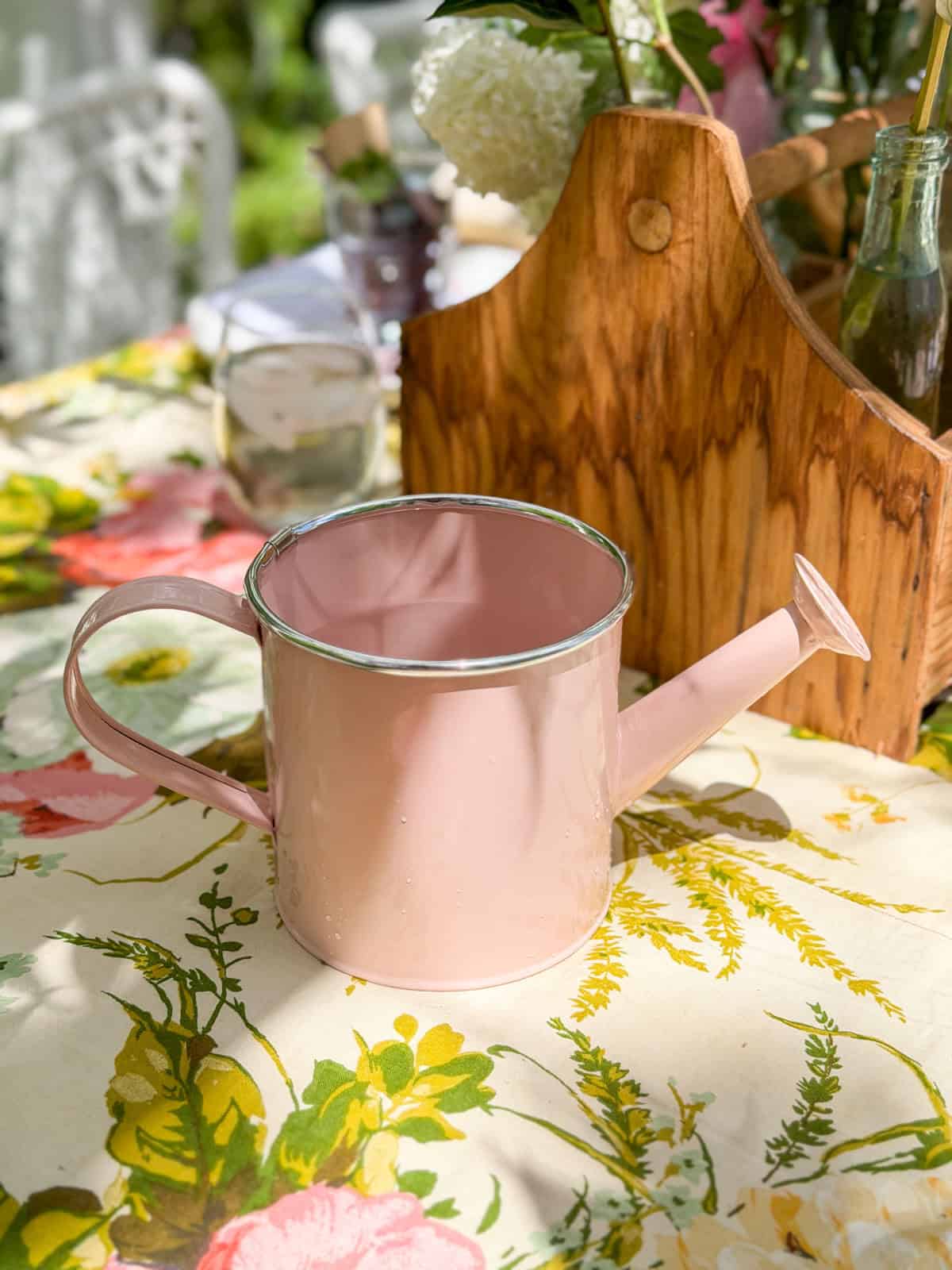 Pink metal watering can used as water pitcher on garden party table beside vintage wooden flower caddy.