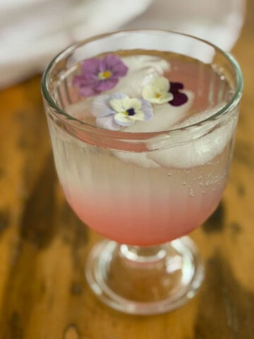 Rhubarb soda in a glass goblet with edible viola flowers floating on ice over pink rhubarb simple syrup.
