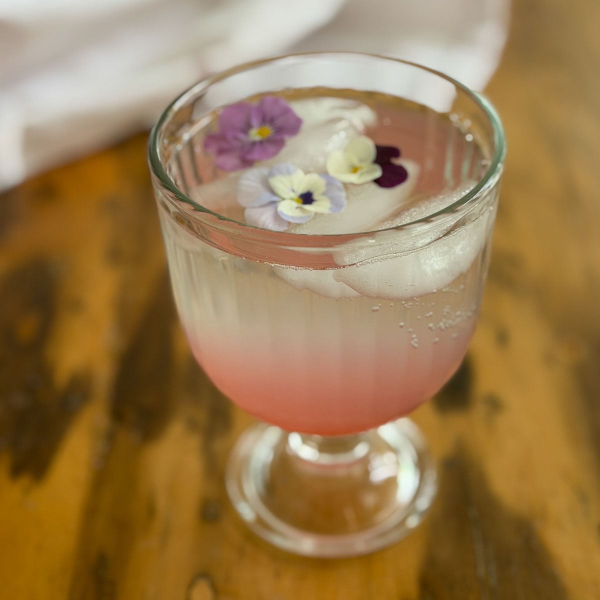 Rhubarb soda in a glass goblet with edible viola flowers floating on ice over pink rhubarb simple syrup.