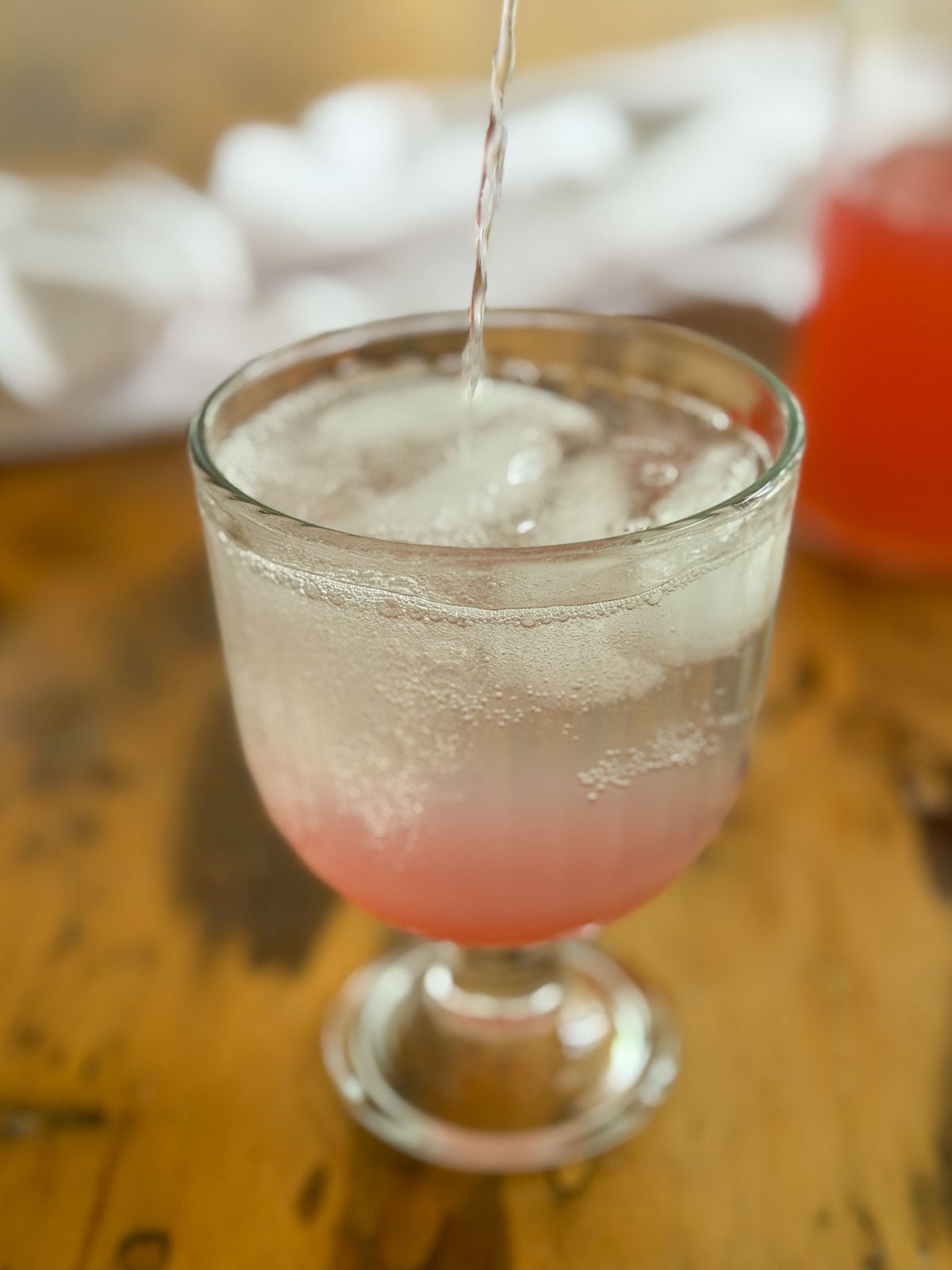 Sparkling water being poured into a glass goblet over ice and rhubarb simple syrup to make homemade rhubarb soda.