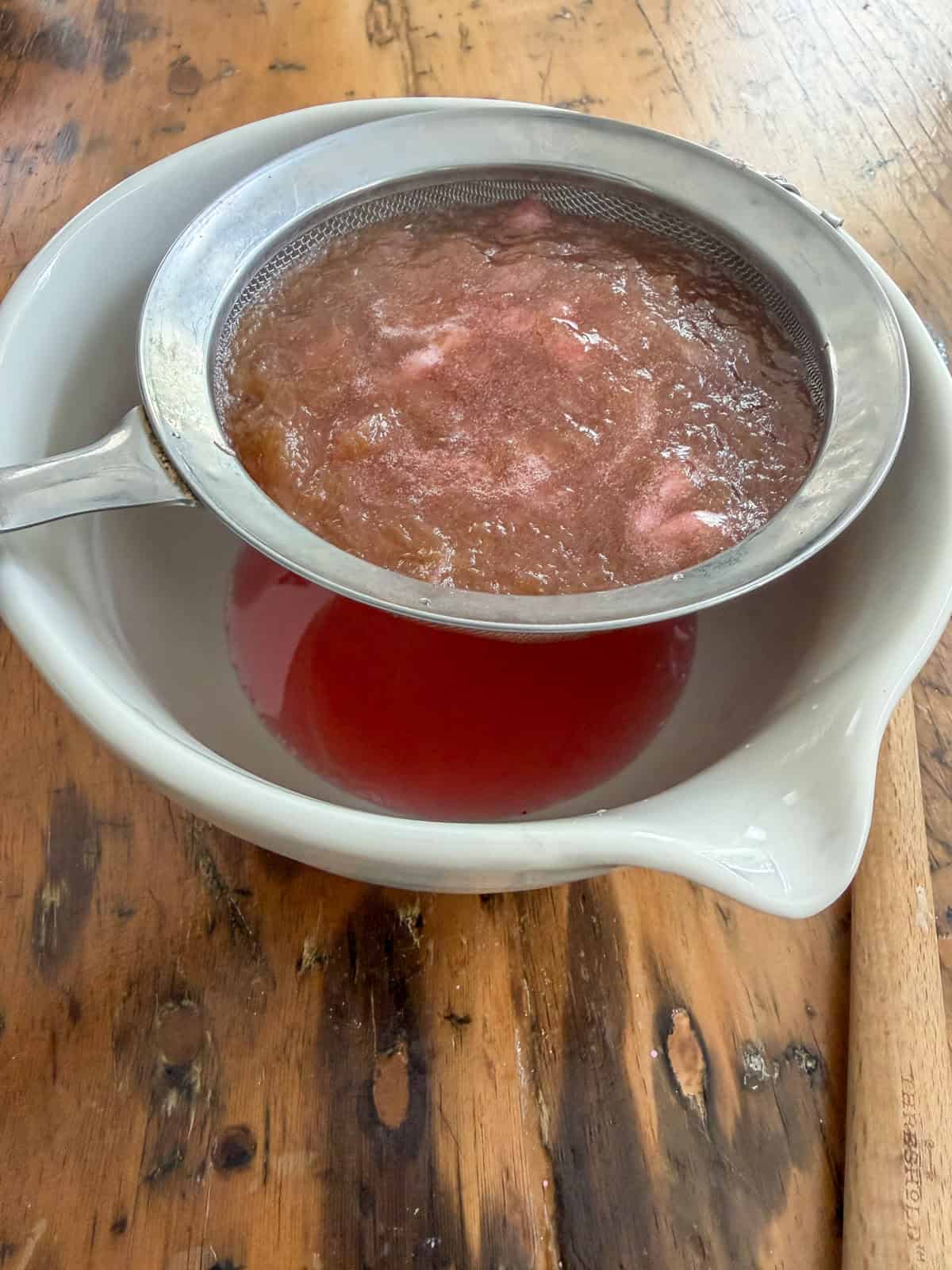 Cooked rhubarb solids in a fine mesh sieve over a white bowl with deep red rhubarb simple syrup draining through.