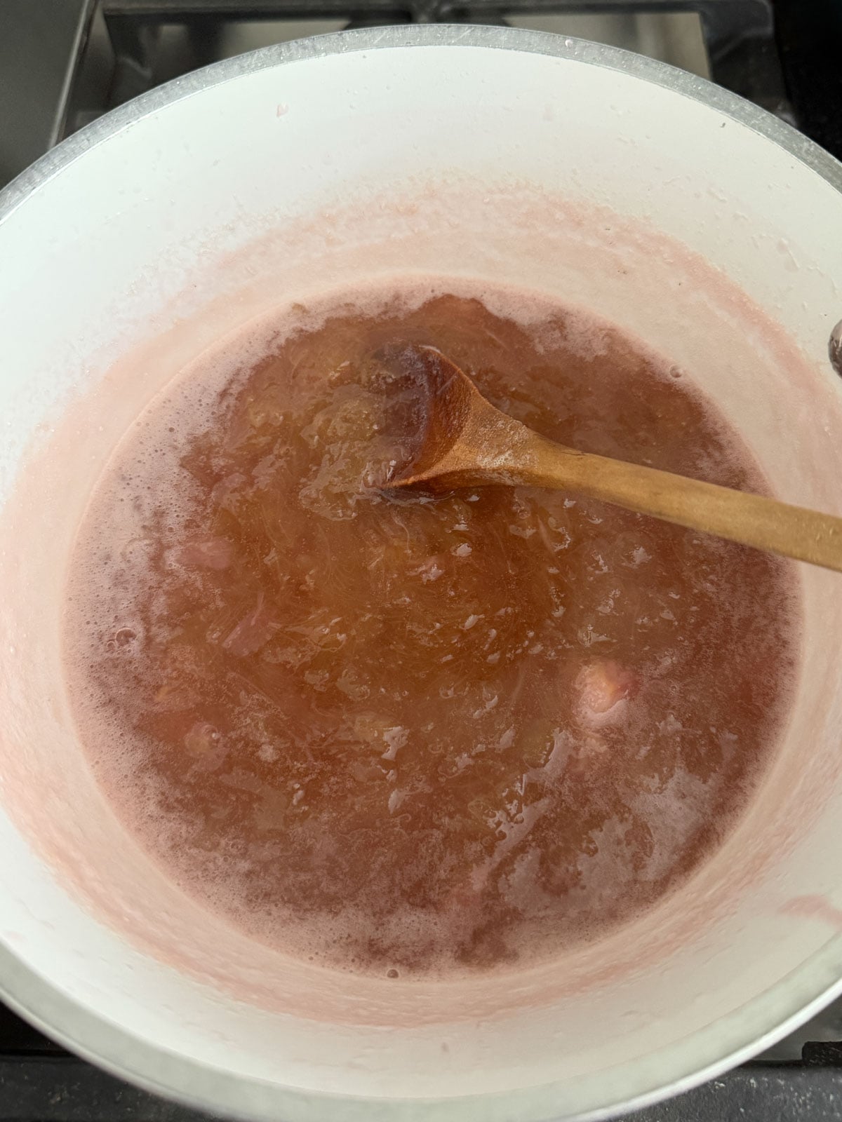 Rhubarb simmering with sugar and water in a white saucepan, breaking down into deep pink syrup while being stirred with a wooden spoon.