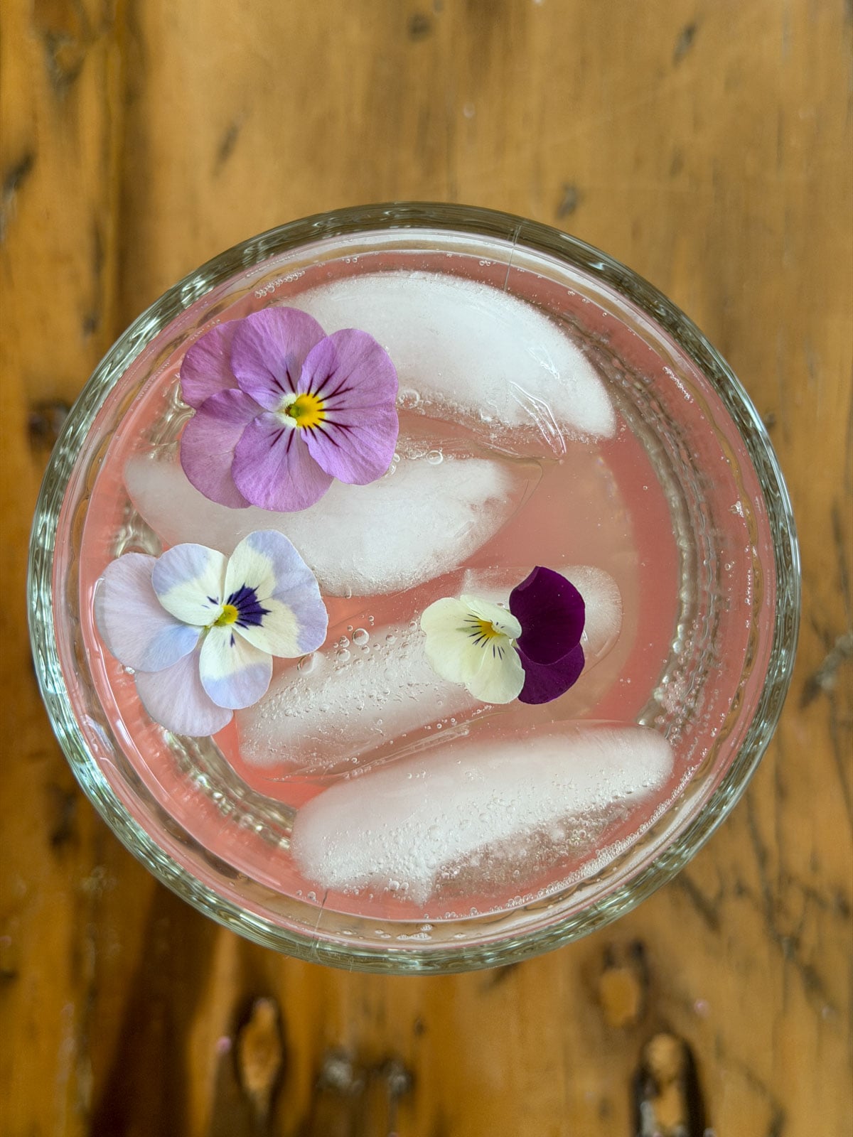 Overhead view of rhubarb soda in a glass goblet with three edible viola flowers floating on pink sparkling water and ice.