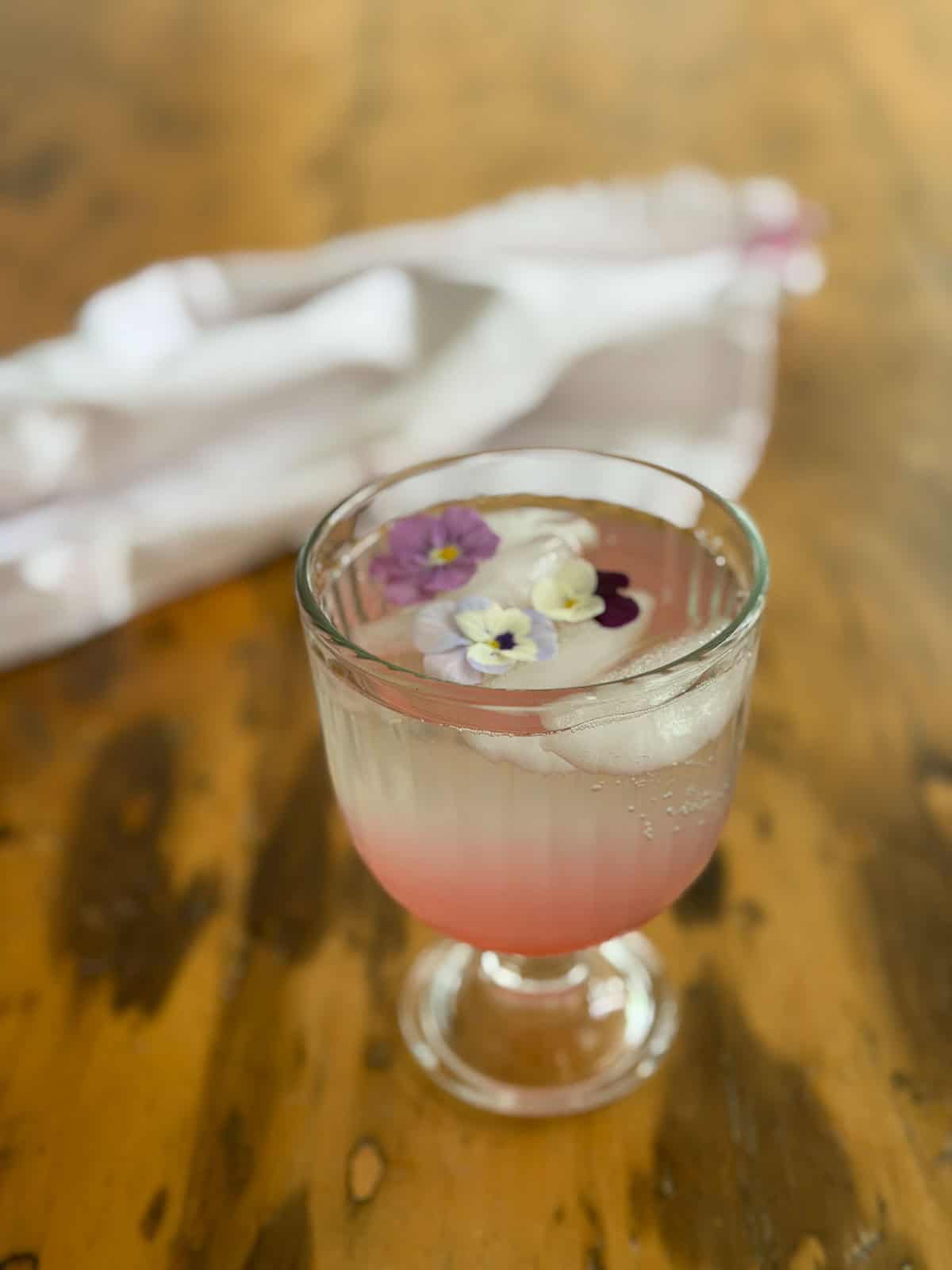 Rhubarb soda in a glass goblet with edible viola flowers floating on ice over pink rhubarb simple syrup.