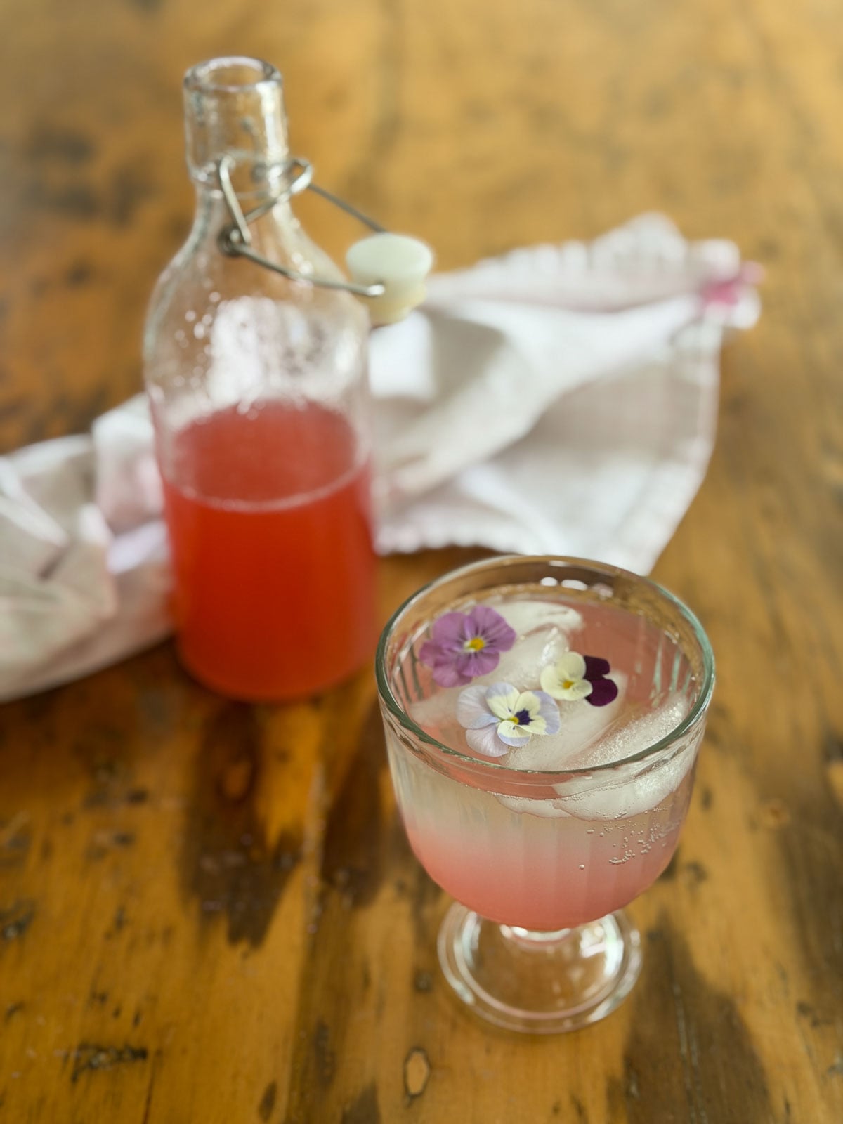 Glass goblet of rhubarb soda with edible flowers beside a swing-top bottle of deep pink rhubarb simple syrup on a wood table.