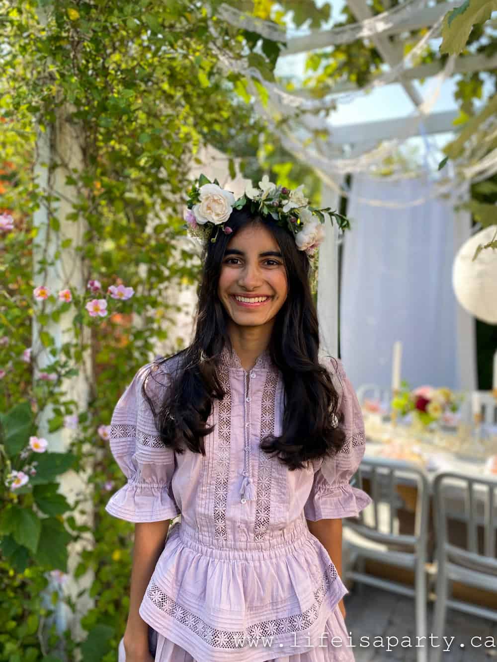 Young woman wearing a handmade garden tea party flower crown of white roses and fresh greenery, smiling in a blooming backyard garden.