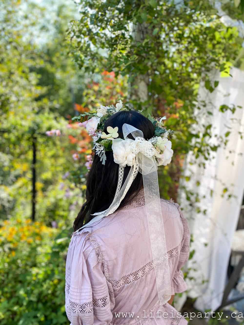 Back view of a handmade garden tea party flower crown with white roses, greenery, and trailing lace ribbon, with colorful autumn garden foliage in background.