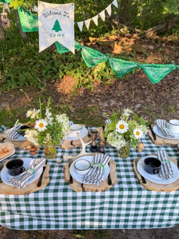 Camp themed party table with green gingham tablecloth, wildflowers, and Welcome to Camp banner.