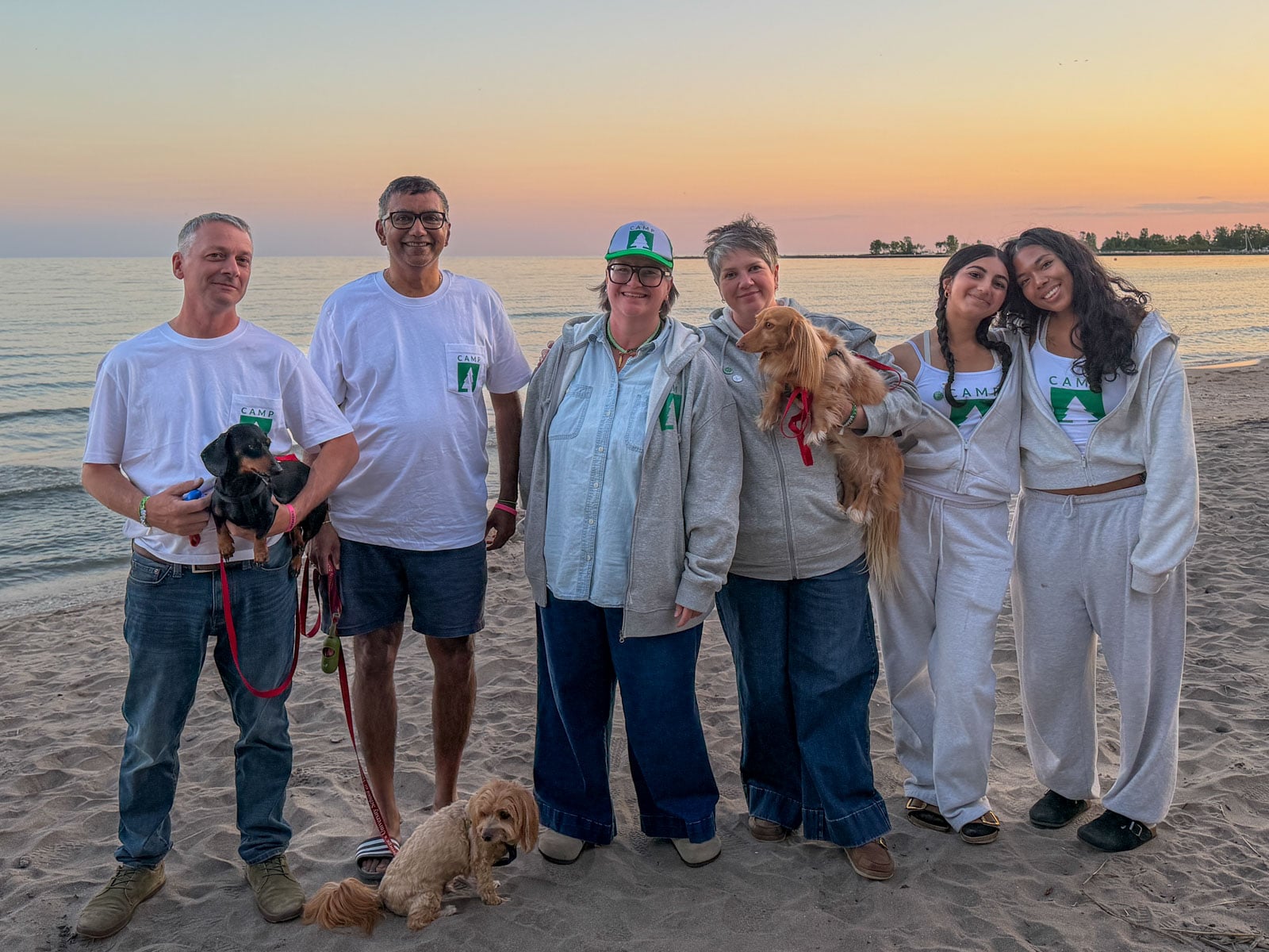 Group wearing Camp logo shirts and hats posing on the beach at sunset with dogs.