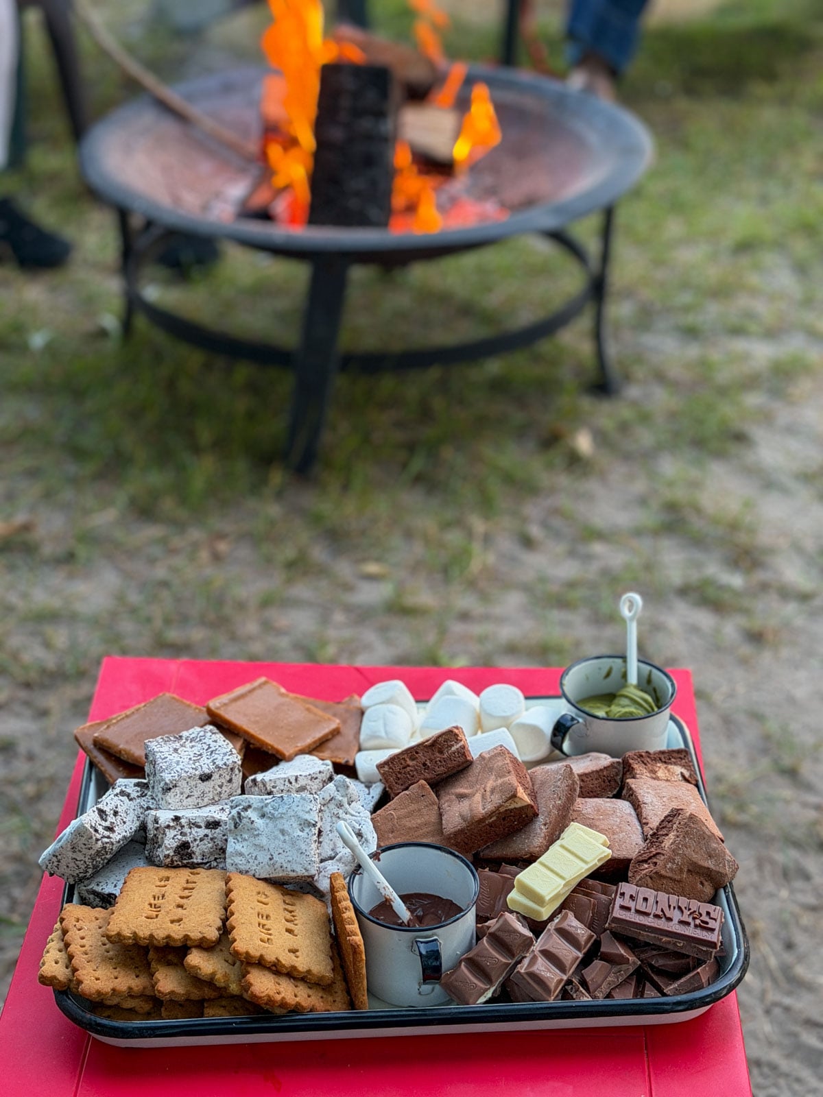 S'mores charcuterie board on red table beside a glowing campfire.