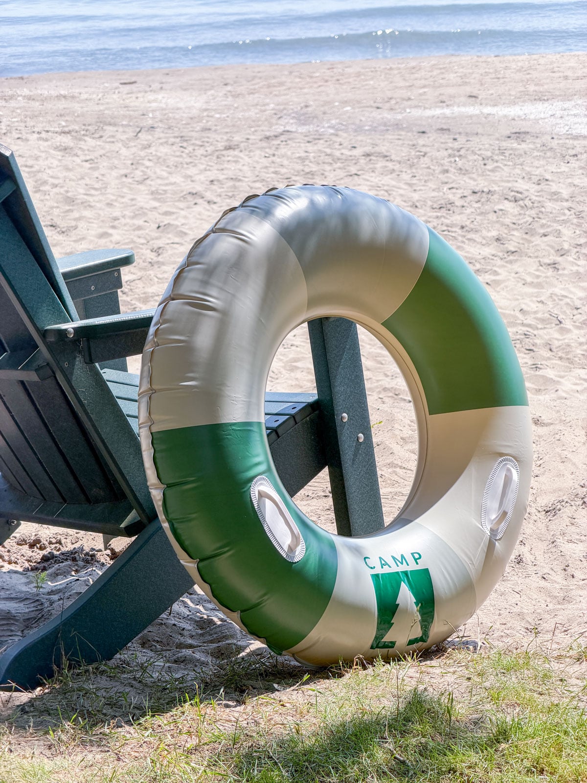 Green and white Camp logo pool float leaning against an Adirondack chair on the beach.