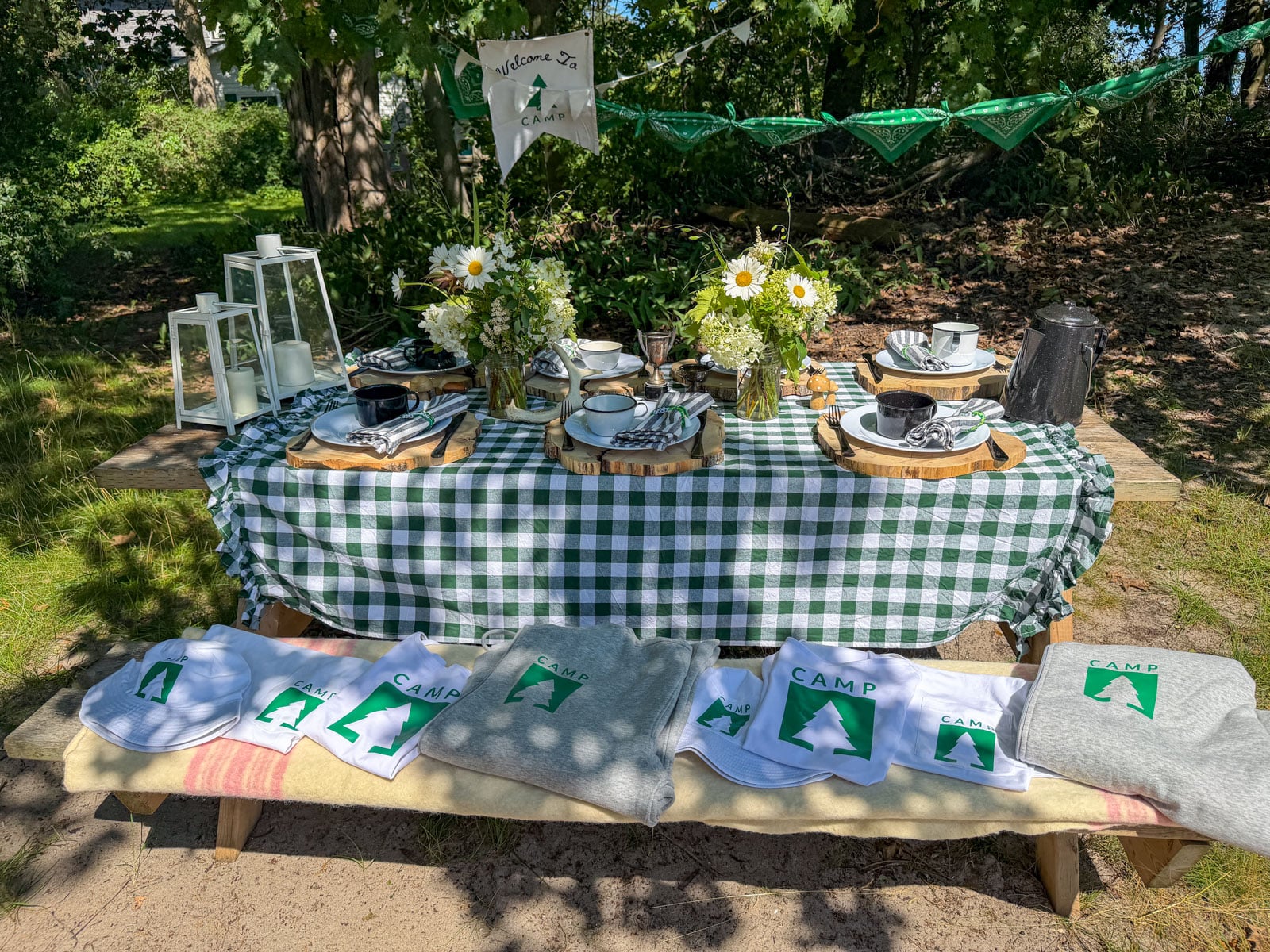 Camp logo t-shirts, hoodies, and hats laid out on picnic bench beside party table.