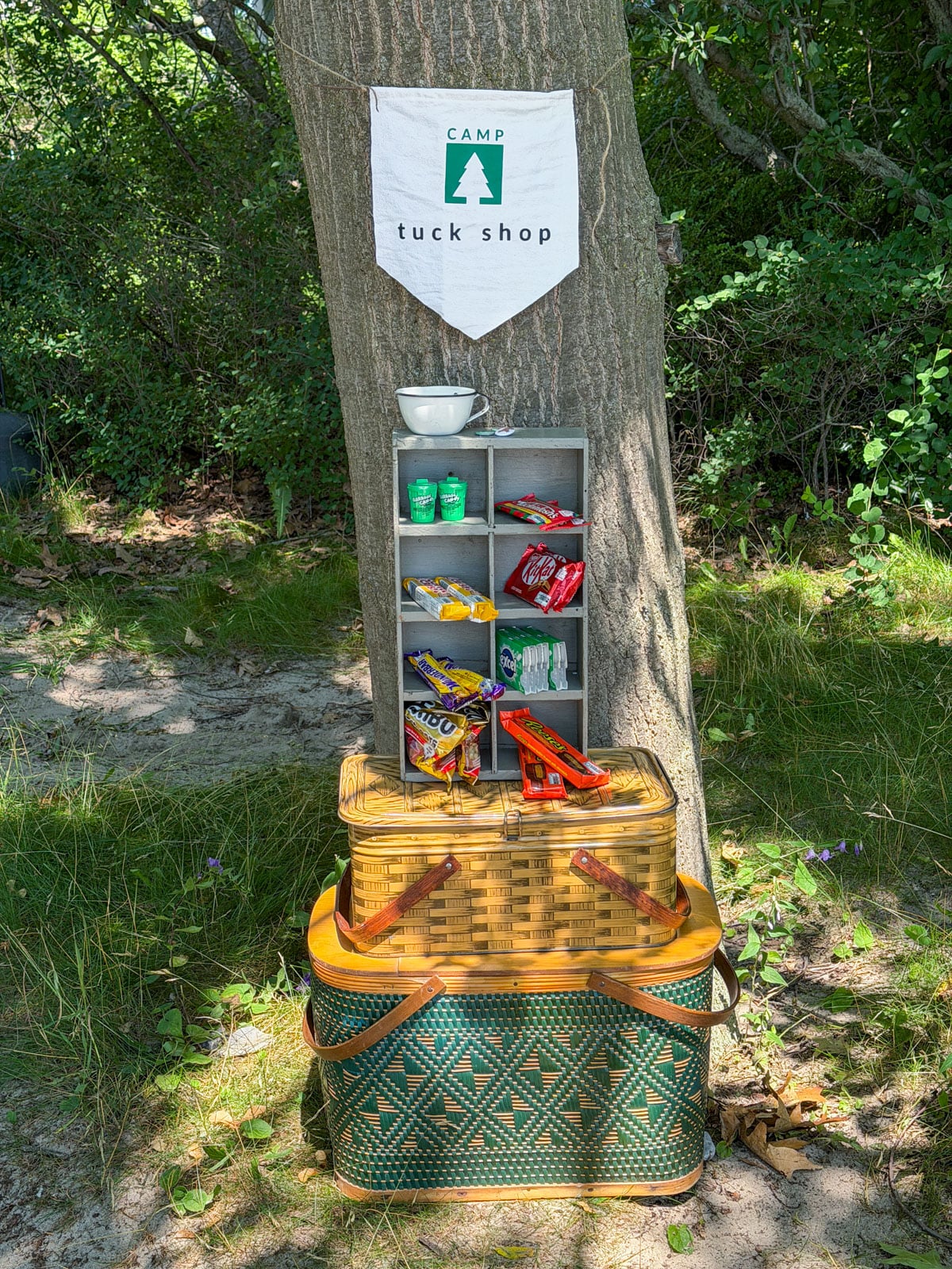 Camp tuck shop display with candy shelf and vintage picnic baskets under a tree.
