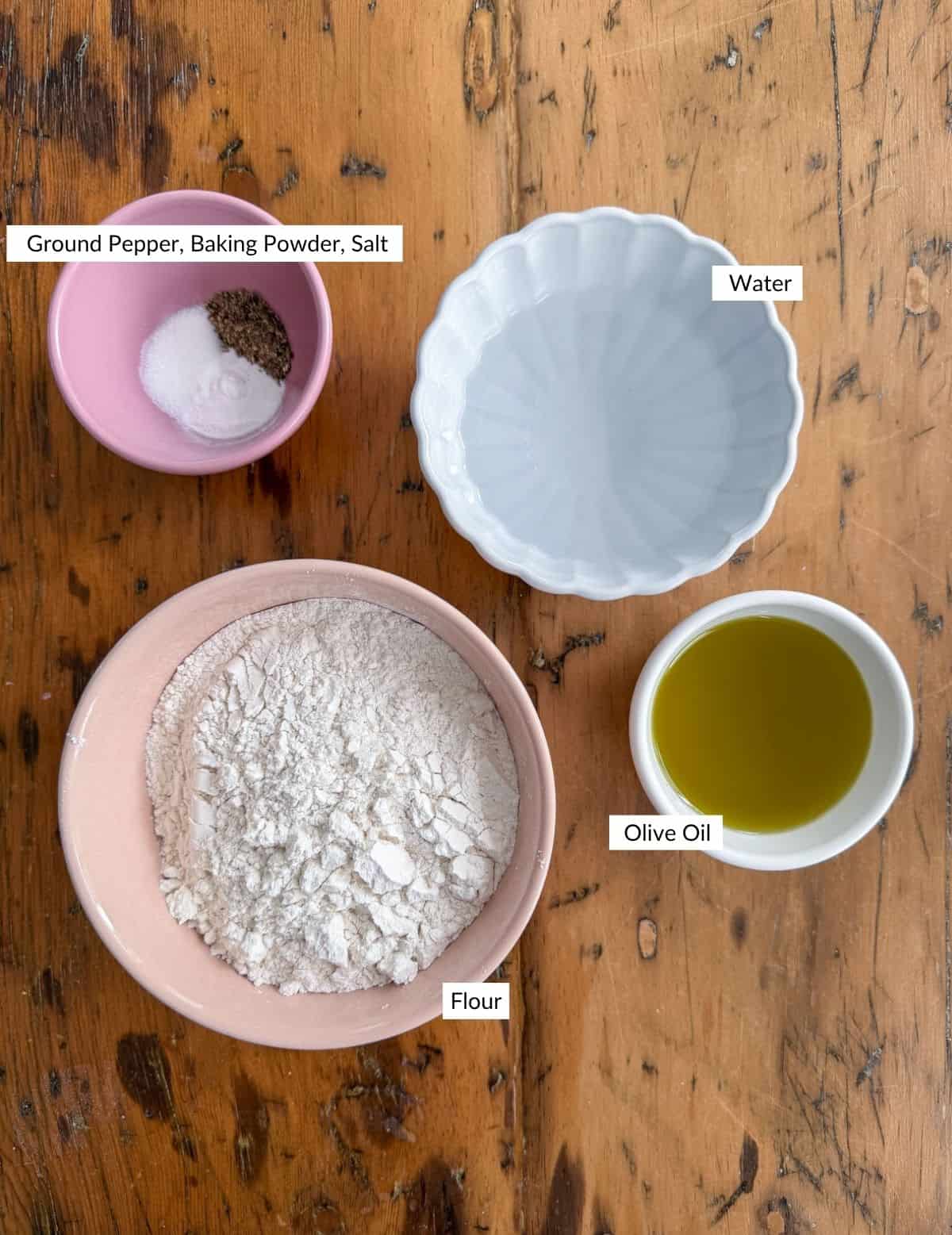 Bowls of flour, olive oil, coarse salt, black pepper, and baking powder measured out for flatbread cracker dough.