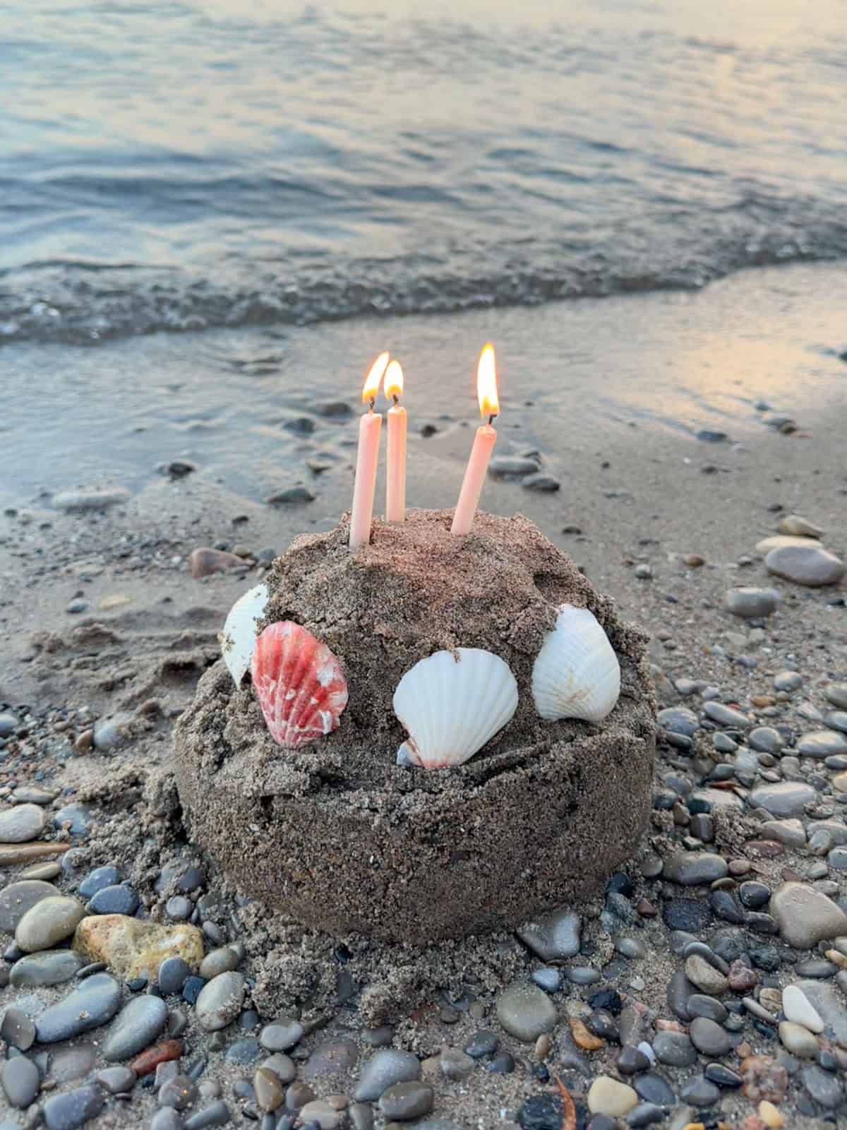 Sand cake decorated with seashells and lit candles on the beach.