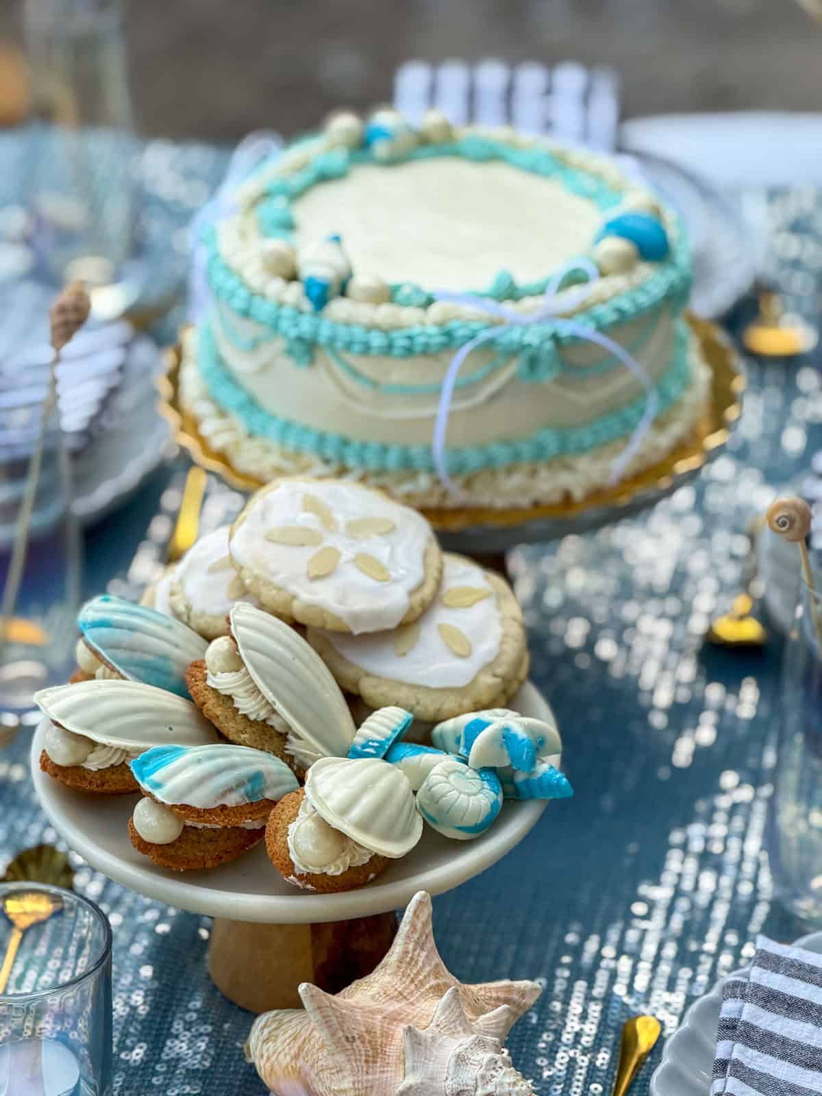 Sand dollar and seashell cookies on a plate at a mermaid party, with a mermaid cake in the background.