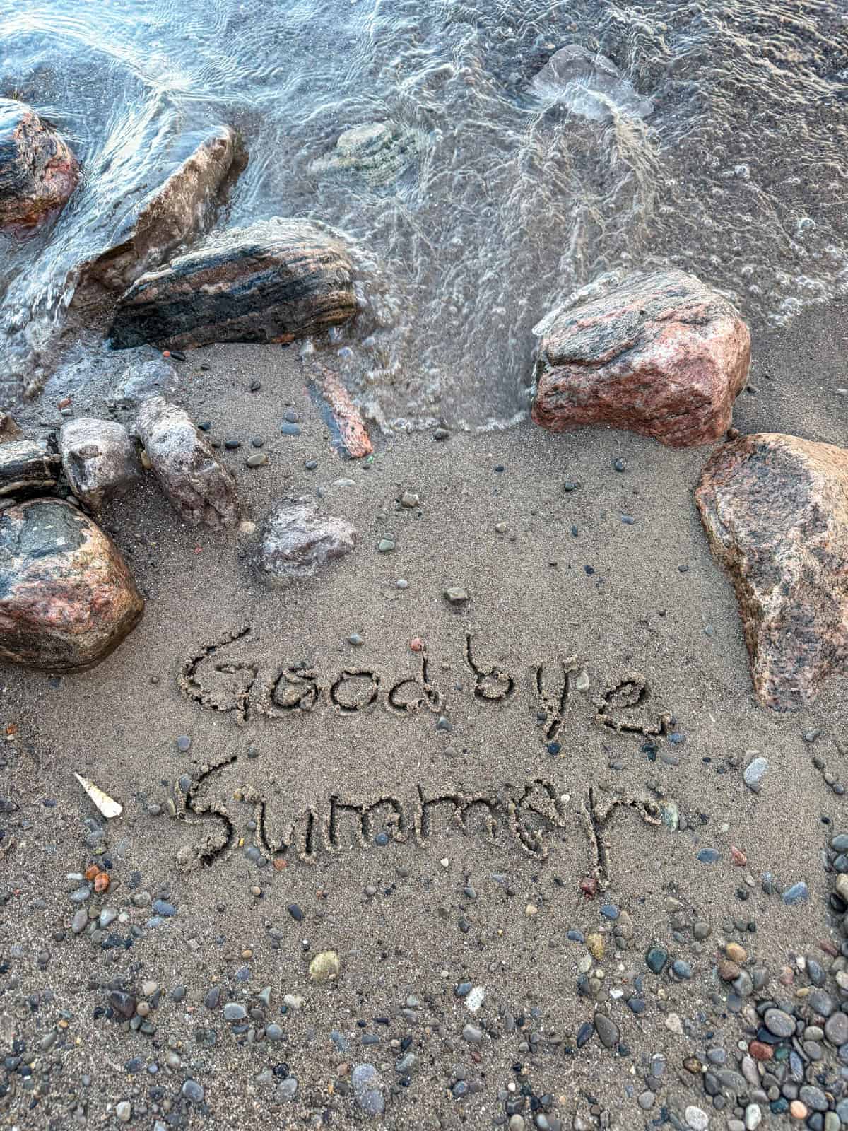 Goodbye Summer written in sand near ocean waves and rocks.