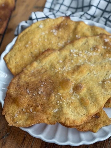 Two golden brown flatbread crackers with cracked pepper and flaky sea salt on a white scalloped plate.