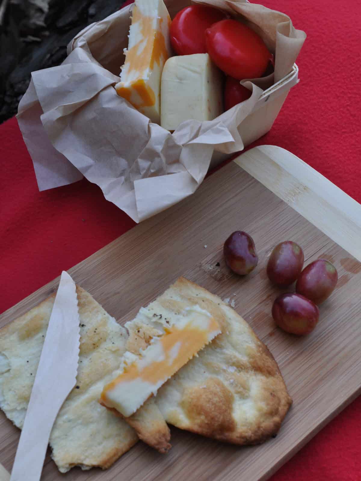 Cubed cheese, cherry tomatoes, red grapes, and flatbread crackers arranged on a wood cutting board.