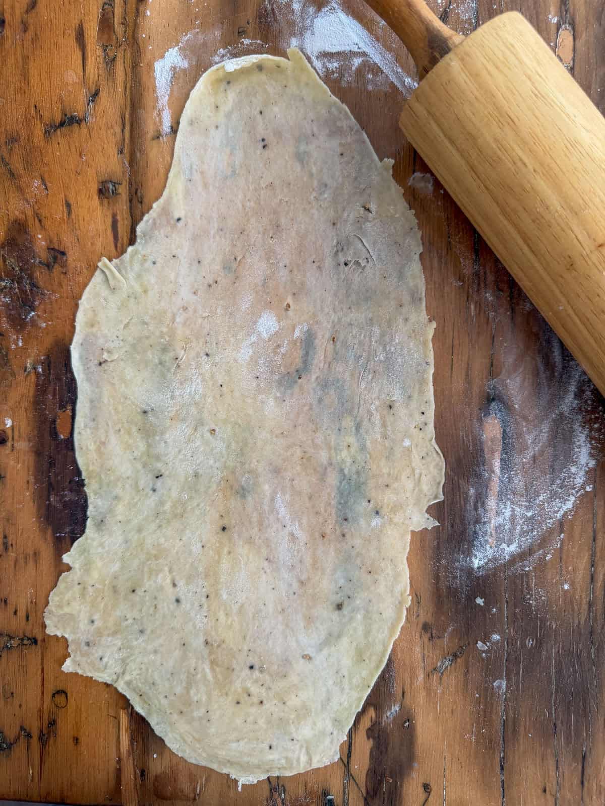 Wooden rolling pin flattening speckled flatbread cracker dough on a floured wood surface.
