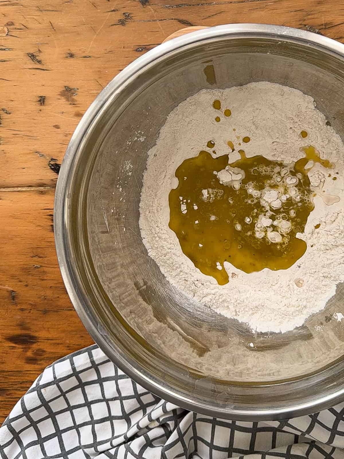 Olive oil poured over flour in a metal mixing bowl for homemade cracker dough.