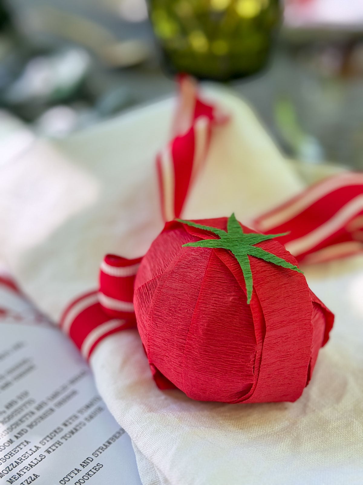 Crepe paper tomato party favor on a white napkin tied with red ribbon.