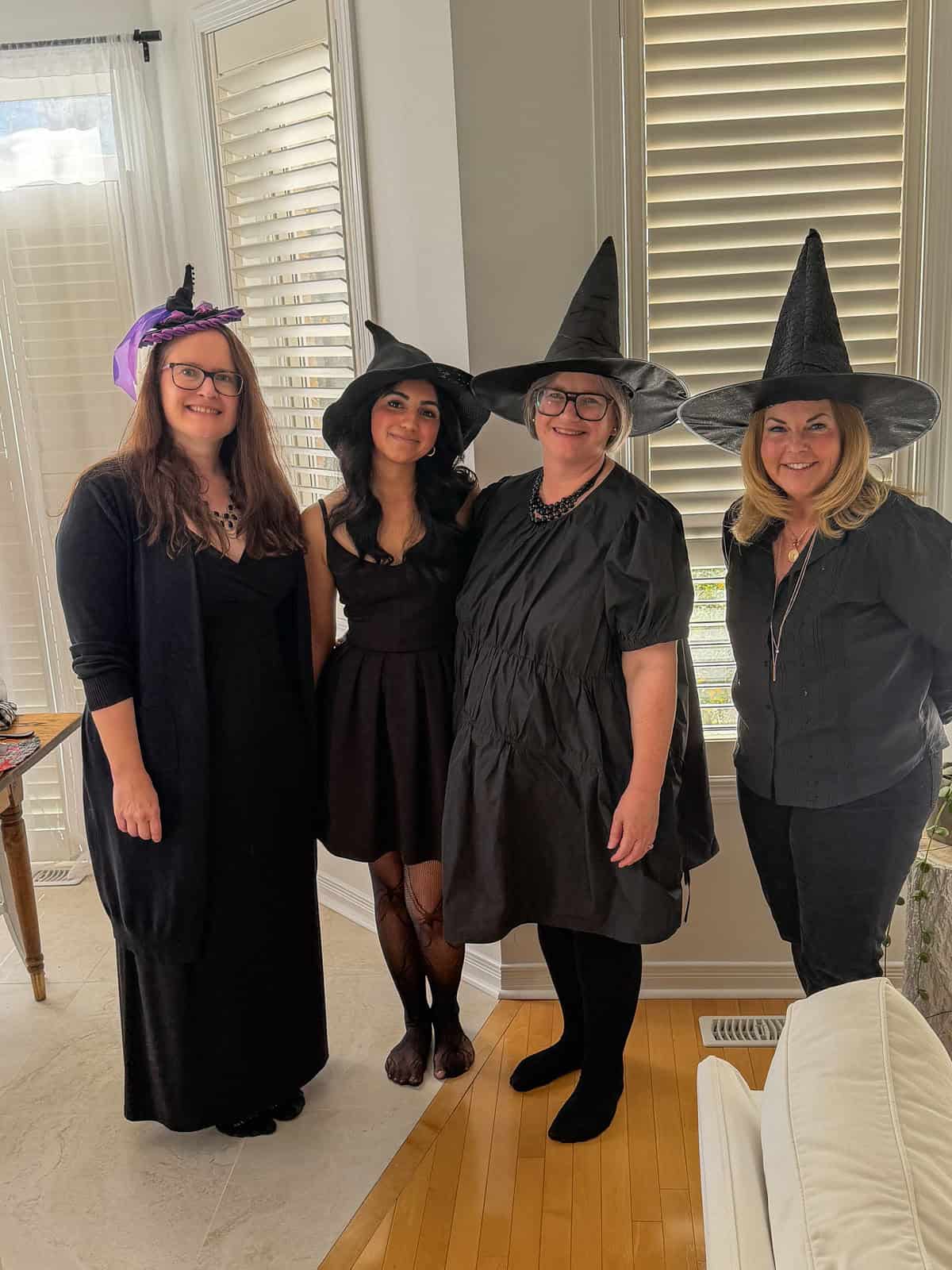 Four friends in black dresses and witch hats standing together in the kitchen.