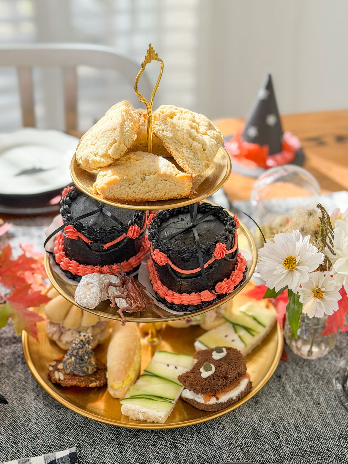 Three tier tray with scones on top, black cakes in the middle and sandwiches on the bottom, with a paper witch hat behind.