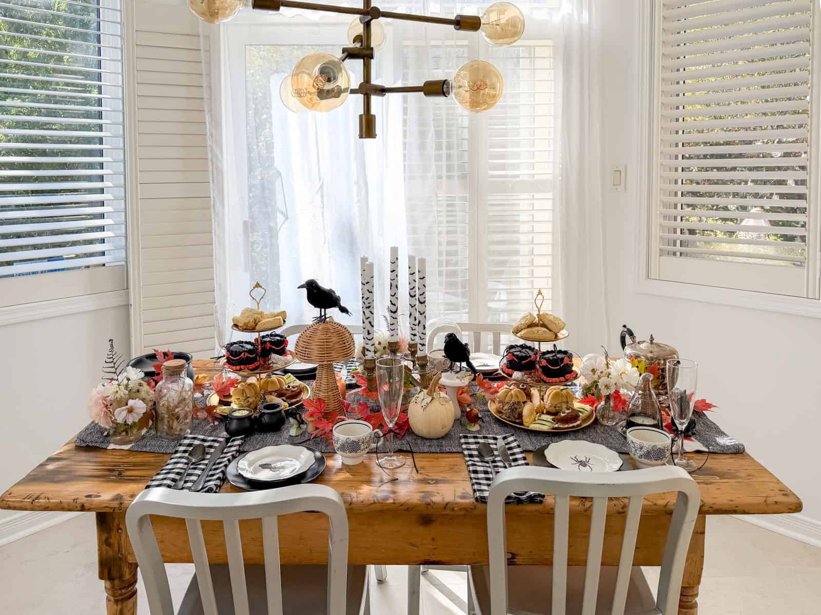 Witch tea party table set for four in a bright kitchen with a gold chandelier above, bat candles and a tiered tray in the center.