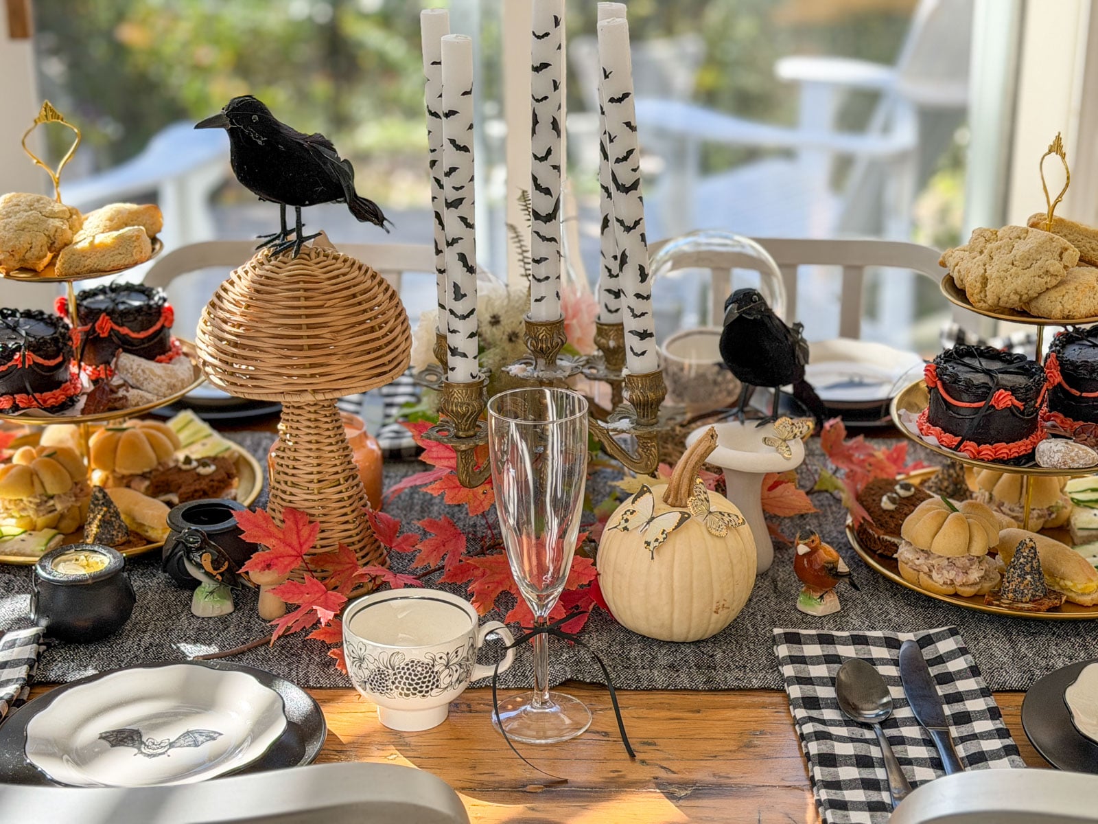 Witch tea party table with a tiered tray on each side of bat candles, a black crow on a wicker mushroom and a white pumpkin.
