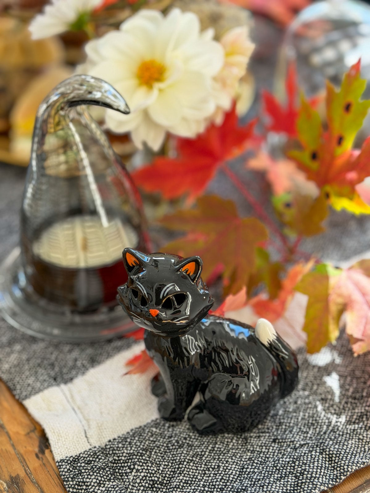Black ceramic cat figurine beside a glass witch hat cloche on a table runner.