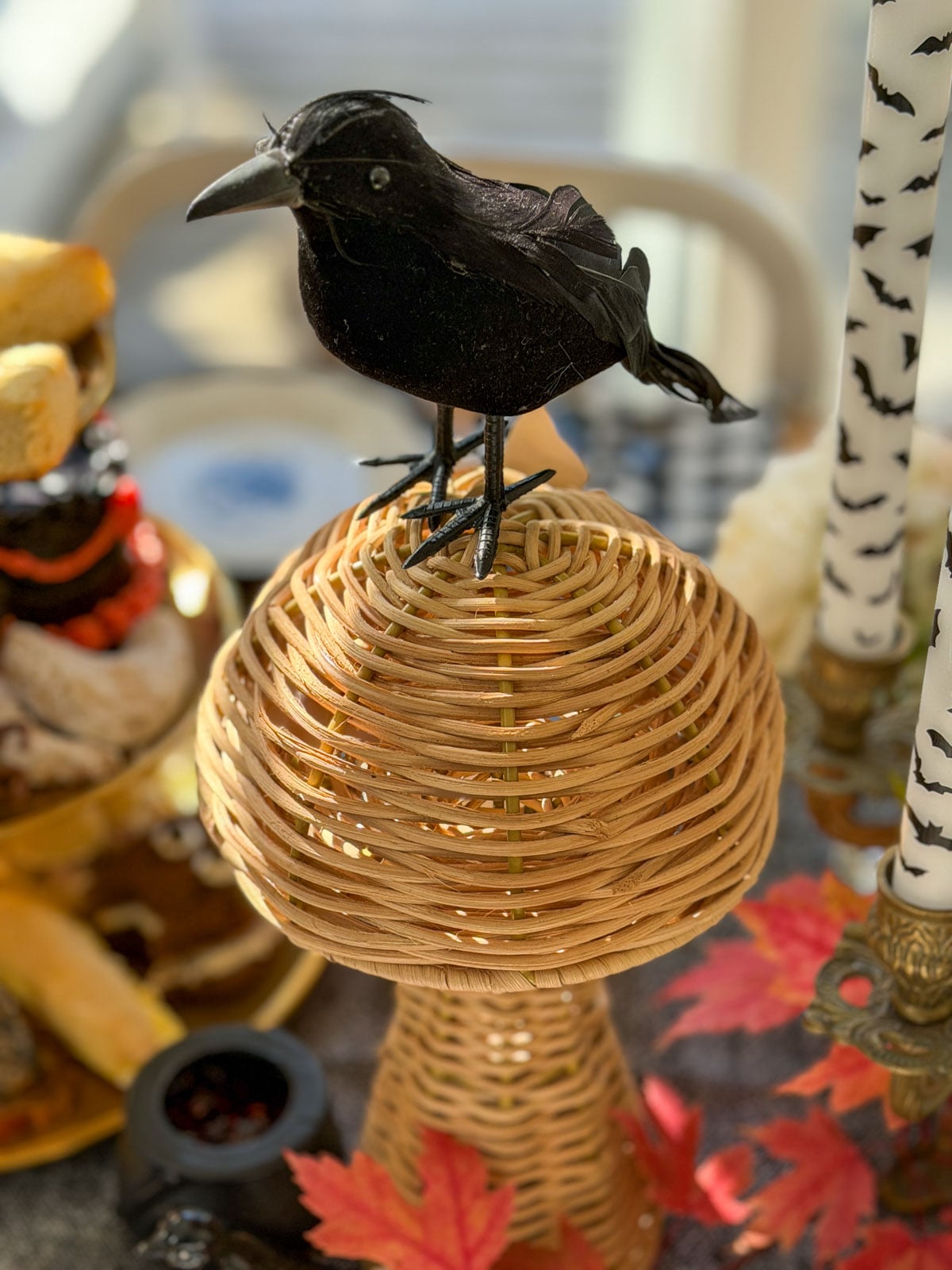 Black crow perched on a wicker mushroom beside bat candles and a tiered tray.