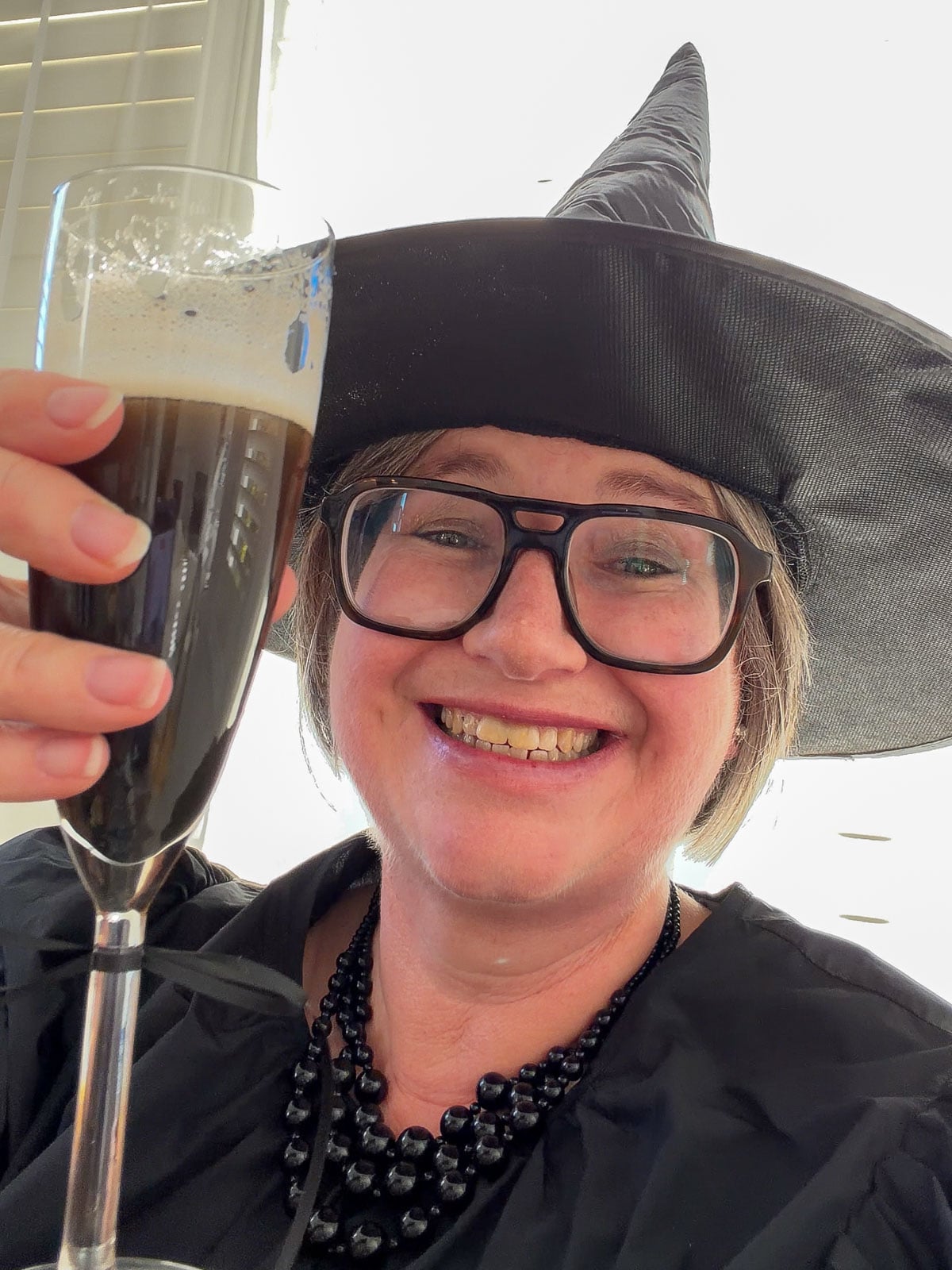 Woman in a black witch hat and black beaded necklace smiling and raising a flute of black Kir Royale.